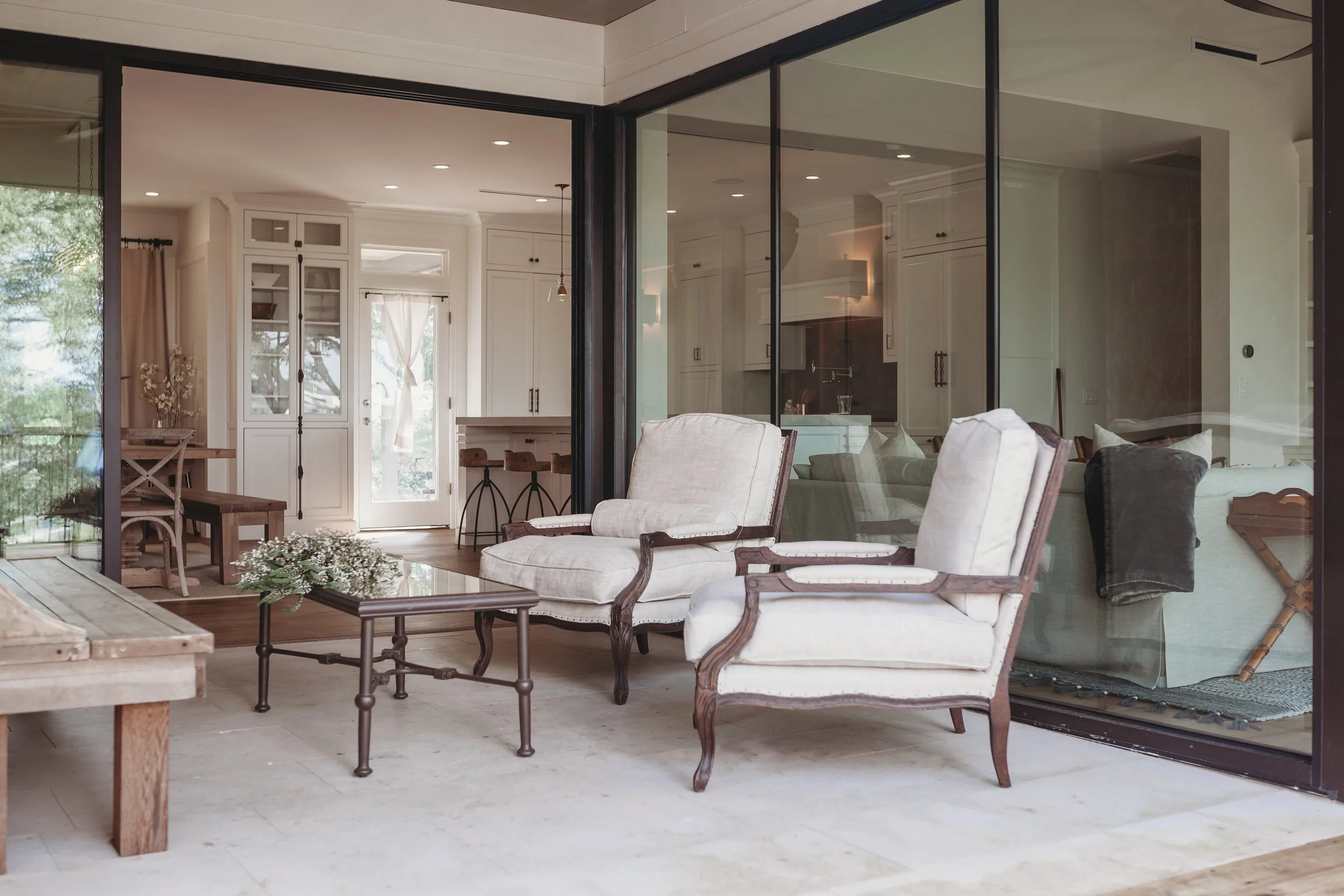Living room with beige armchairs, wooden coffee table, glass sliding doors, and kitchen area with white cabinets.