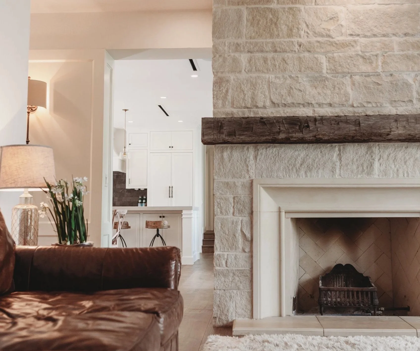 Living room with stone fireplace, wooden mantel, leather sofa in foreground, white kitchen cabinets in background, and a vase with flowers on a side table.