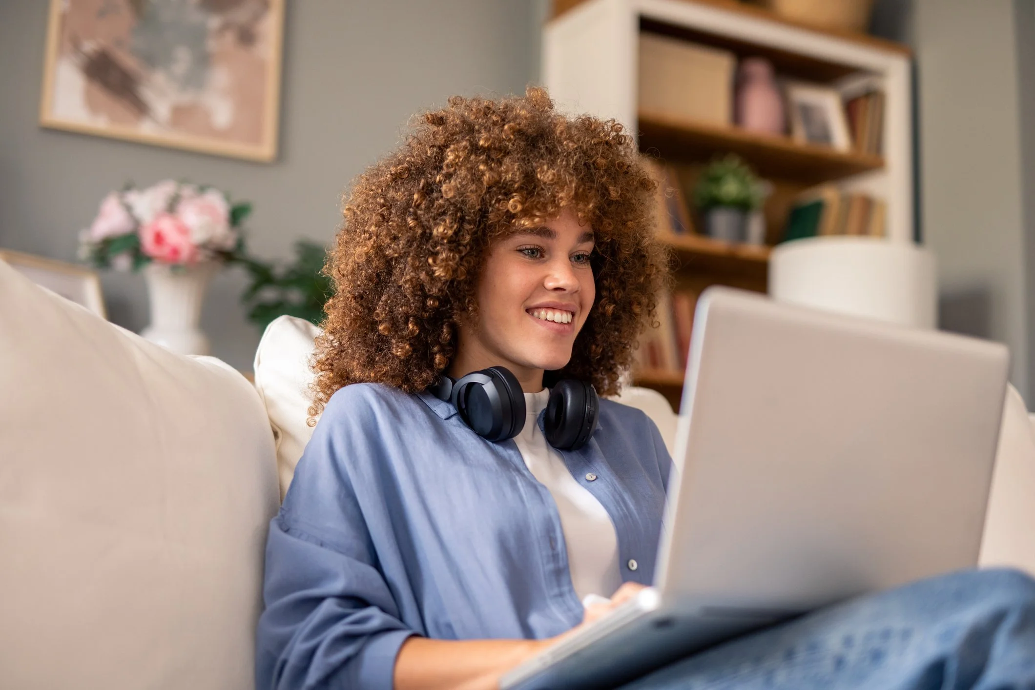 A young woman with curly hair wearing headphones, smiling while looking at her laptop, sitting on a couch with a blurred background of a bookshelf and a flower vase.