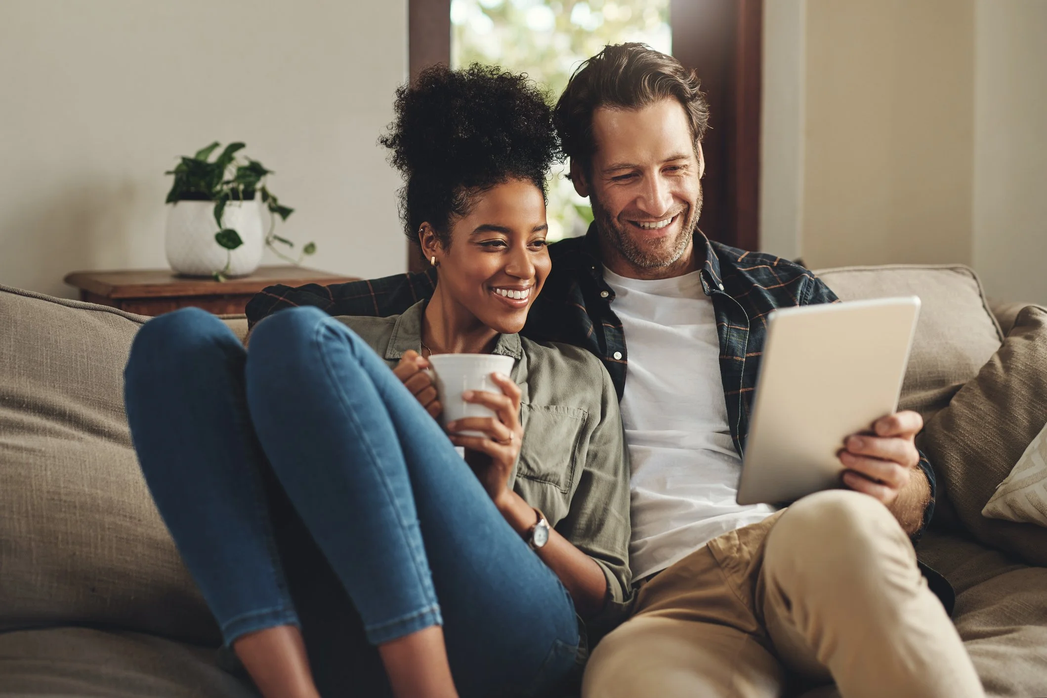 A smiling couple sitting close together on a beige couch, looking at a tablet device. The woman is holding a white mug, and the man is wearing a dark plaid shirt. A potted plant is visible on a wooden table behind them, with a bright window in the background.