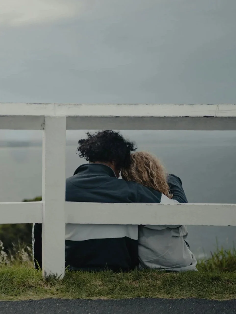 A couple sitting on the grass, viewed from behind, leaning into each other under a cloudy sky with a white fence in the foreground.