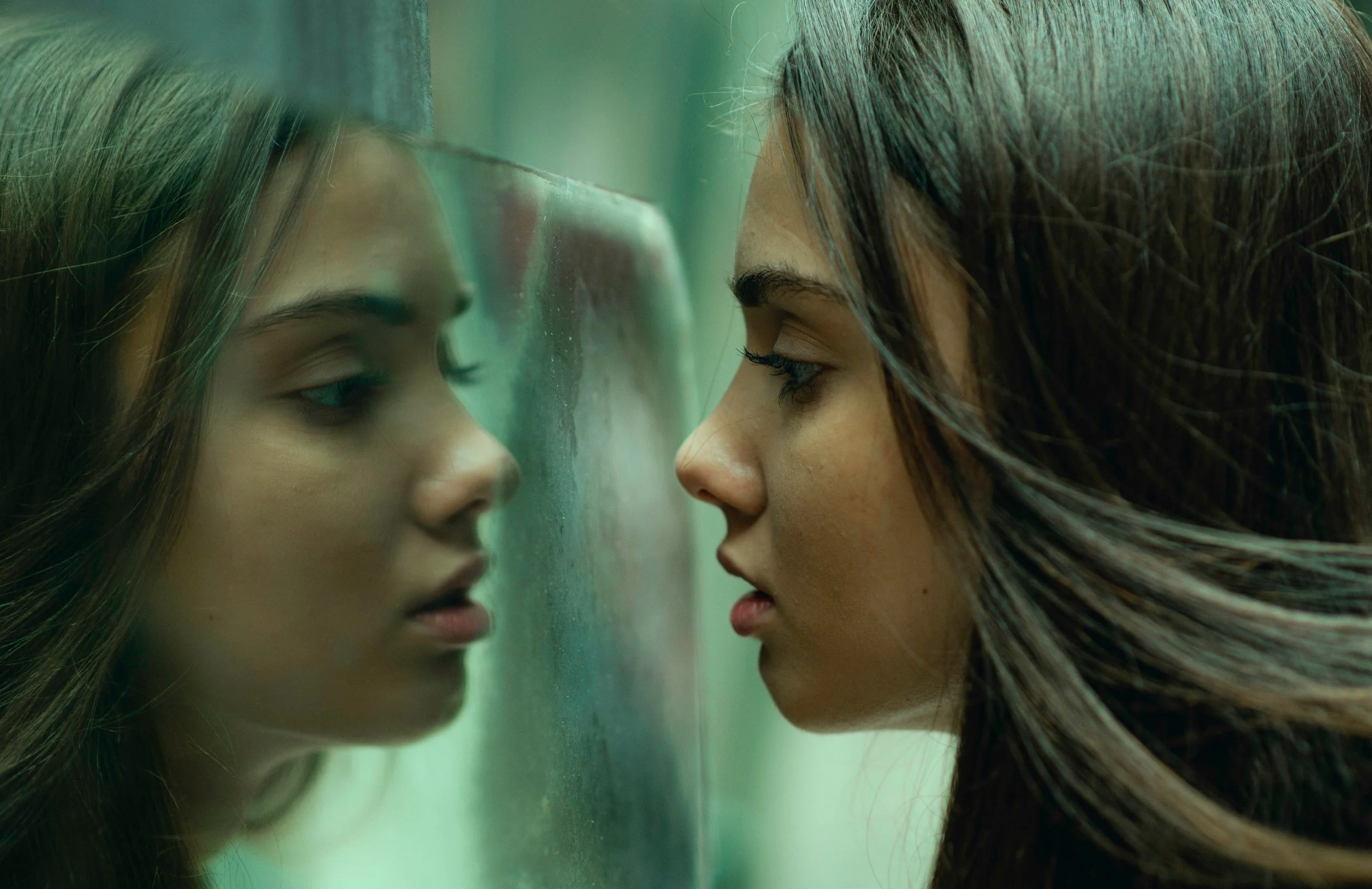 A young woman with long hair looking at her reflection in a glass surface, with her face close to the glass, creating a mirrored image.