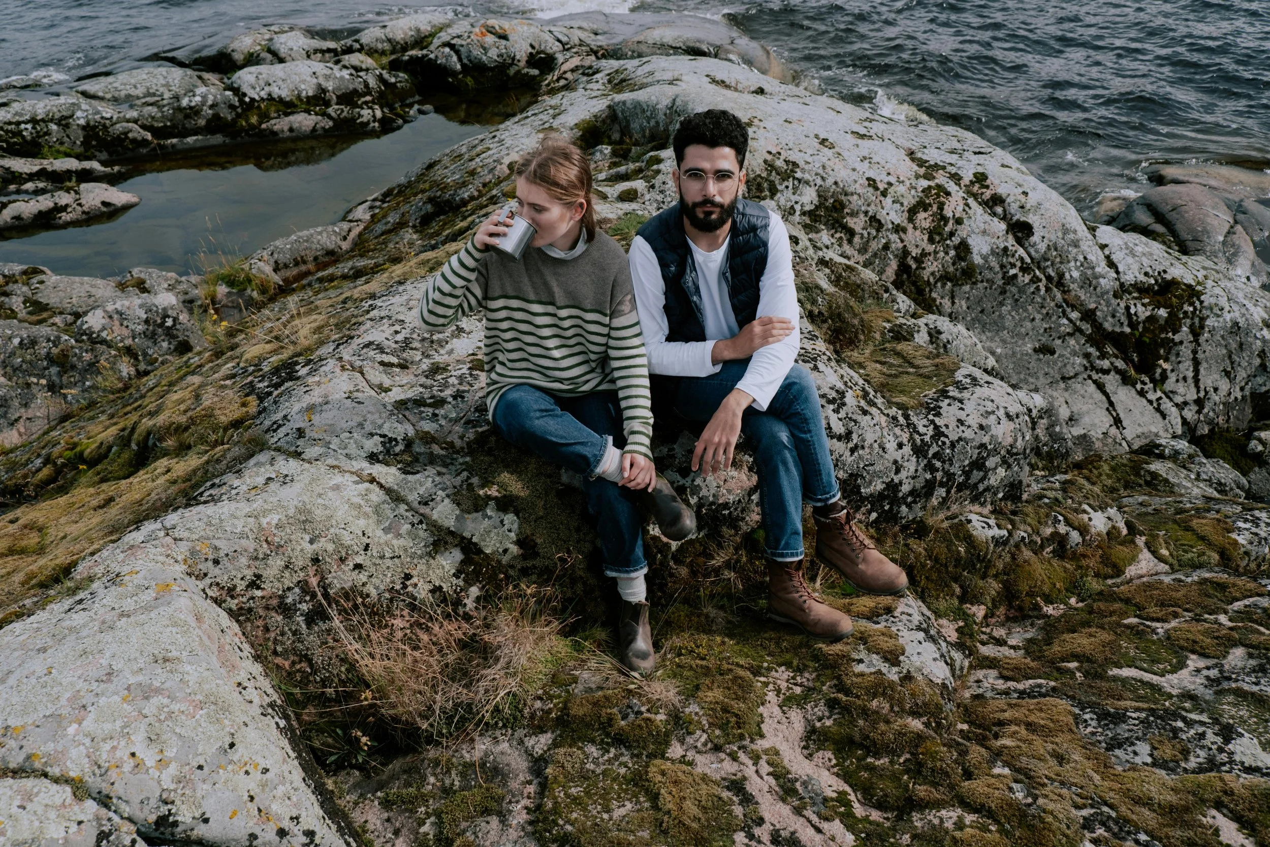 Two people sitting on large rocks near the water. The woman is drinking from a metal cup, and the man is looking at the camera. They are dressed casually in outdoor clothing.