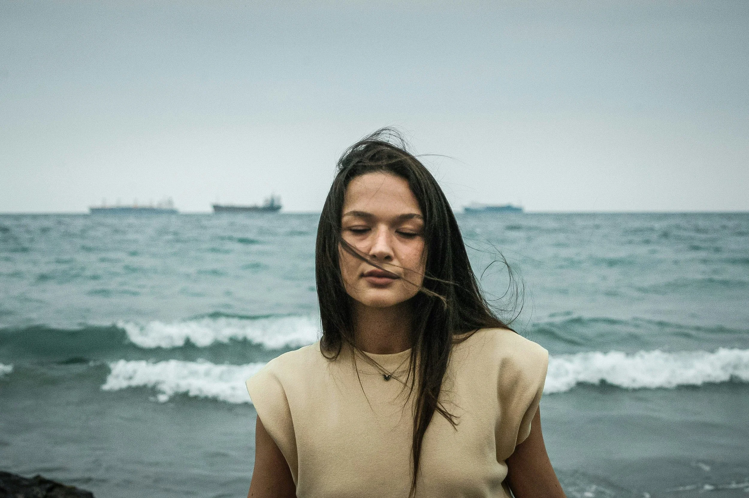 A woman with long dark hair standing in front of the ocean with ships in the background, eyes closed.