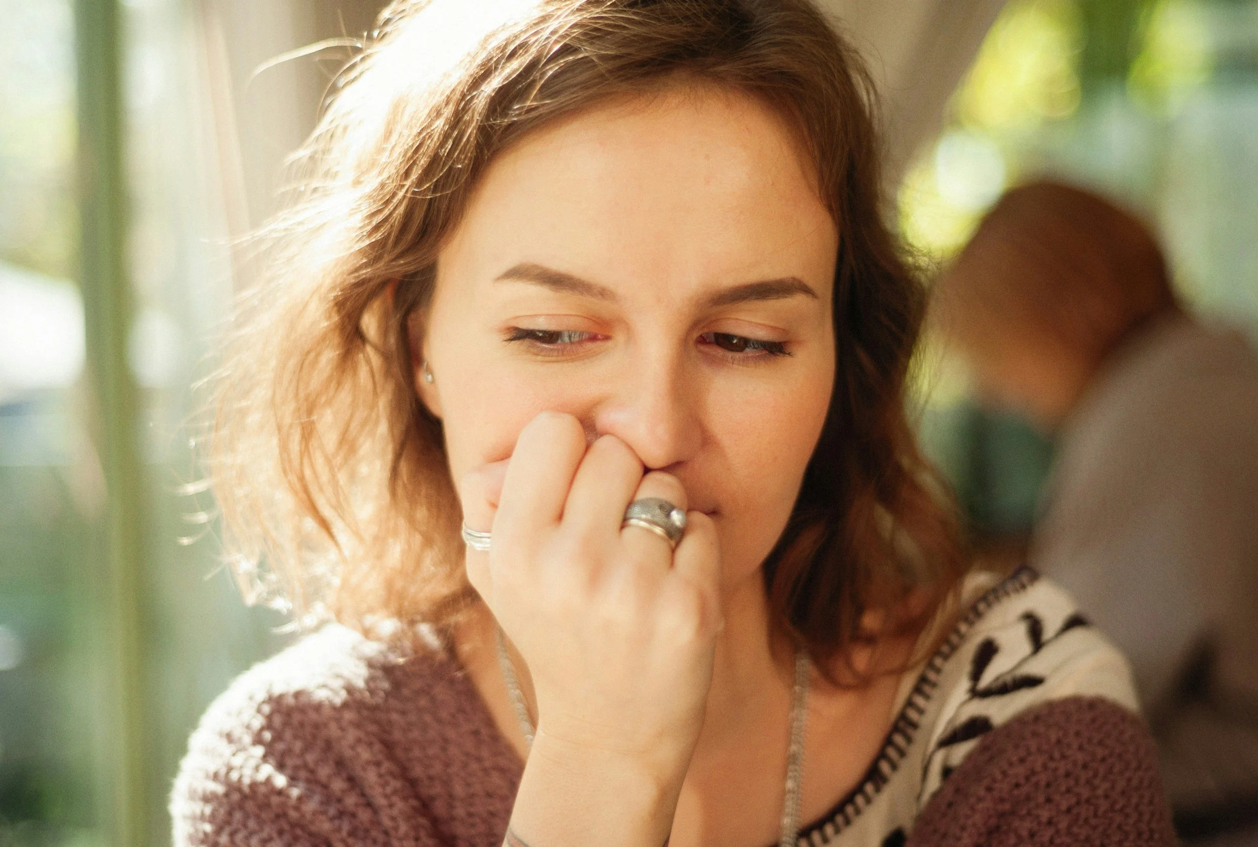 A young woman with light brown, curly hair, wearing a striped shirt, looking down and touching her face with her hand, in a softly lit environment with a blurred green background.