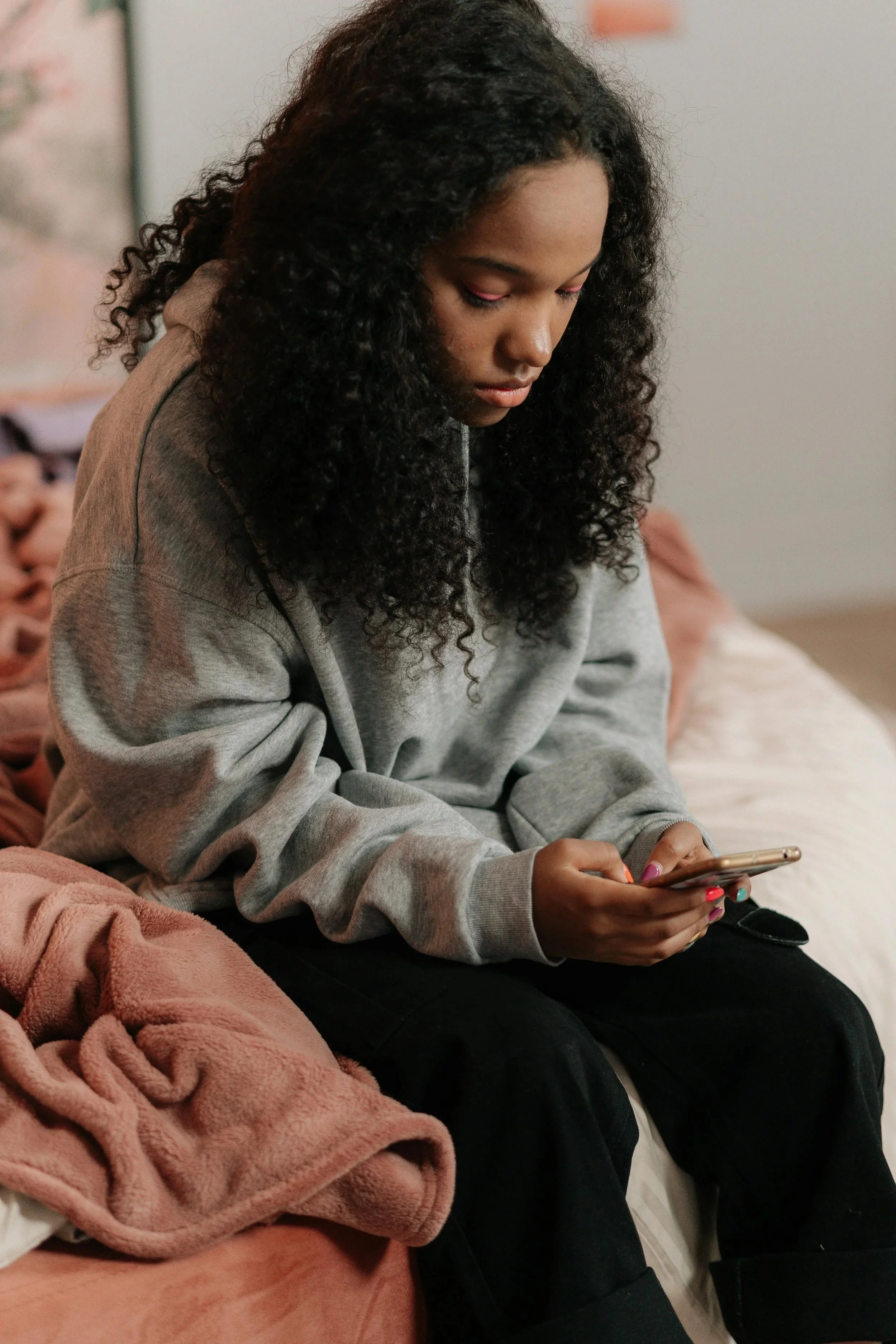 A young woman with curly black hair, wearing a gray hoodie, sitting on a couch and looking at her phone.