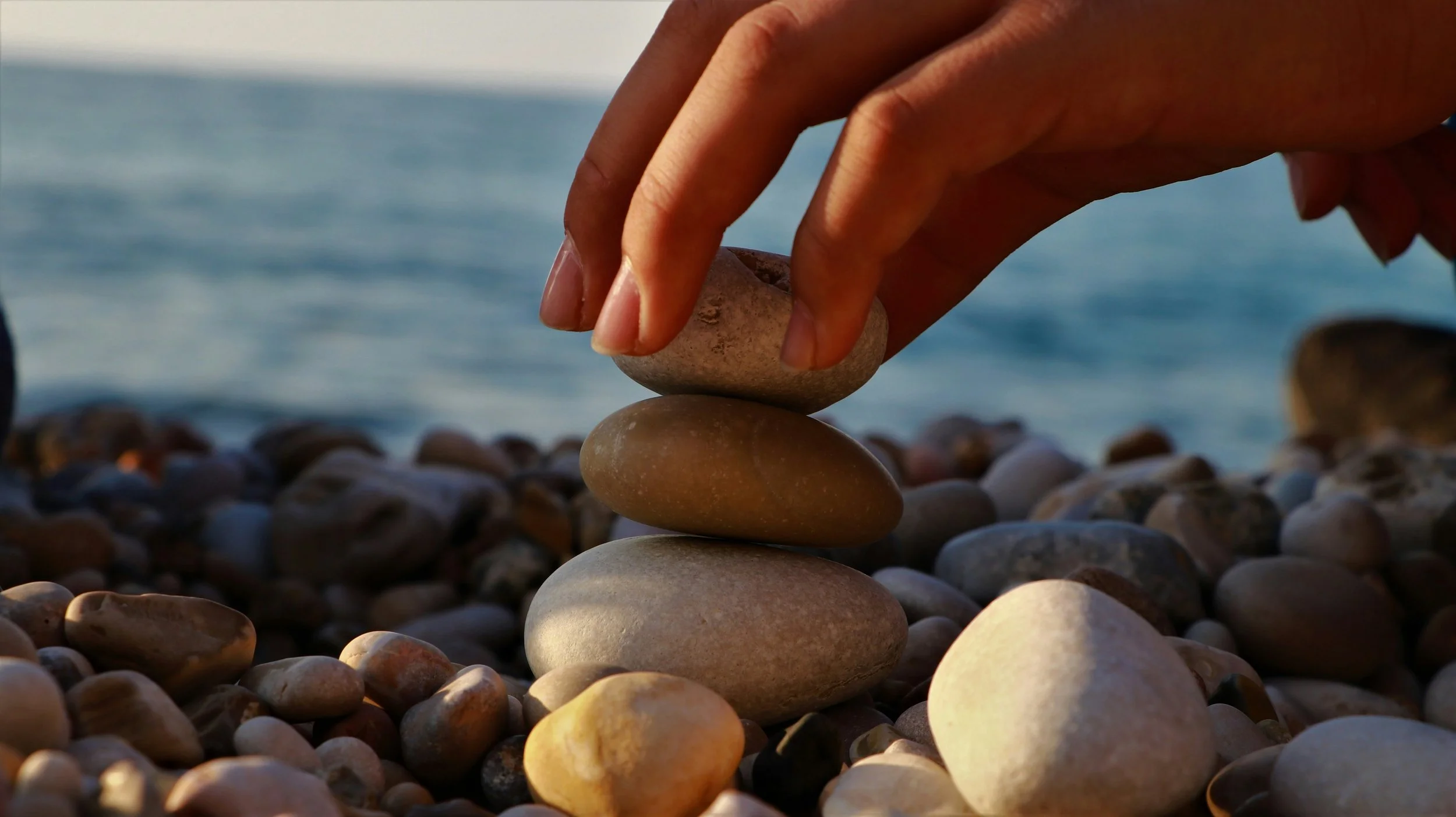 A hand stacking smooth, rounded rocks on a pebble beach near the ocean, with more rocks and water in the background.