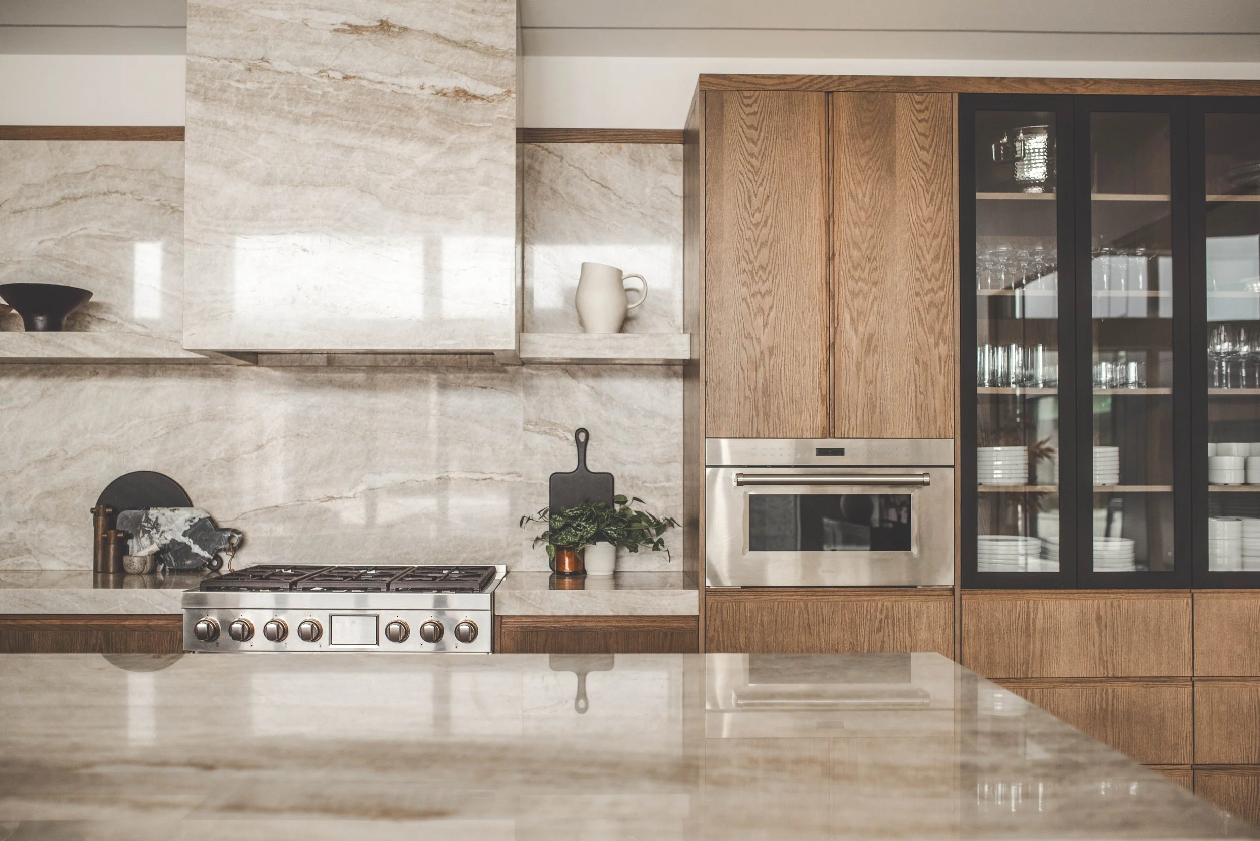 A modern kitchen with beige marble backsplash, wooden cabinetry, stainless steel oven and stove, black glass cabinet doors, and various kitchen items including a white pitcher and a potted plant.