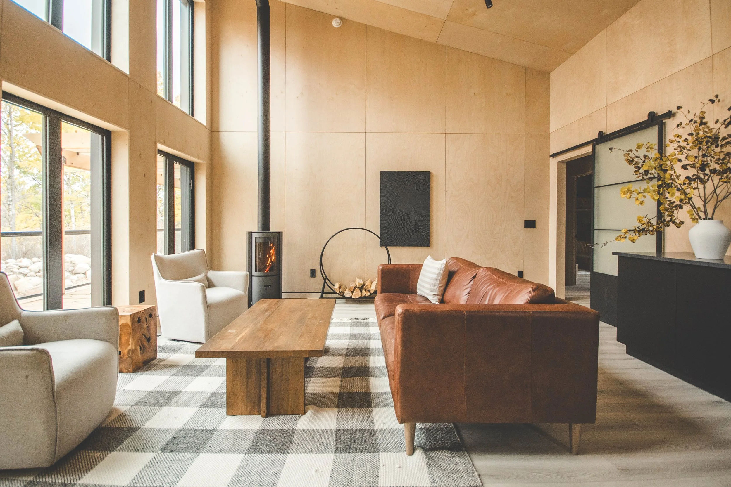 Modern living room with large windows, a wood-burning fireplace, a brown leather sofa, white armchairs, a wooden coffee table, and a black sideboard with a white vase and plant.