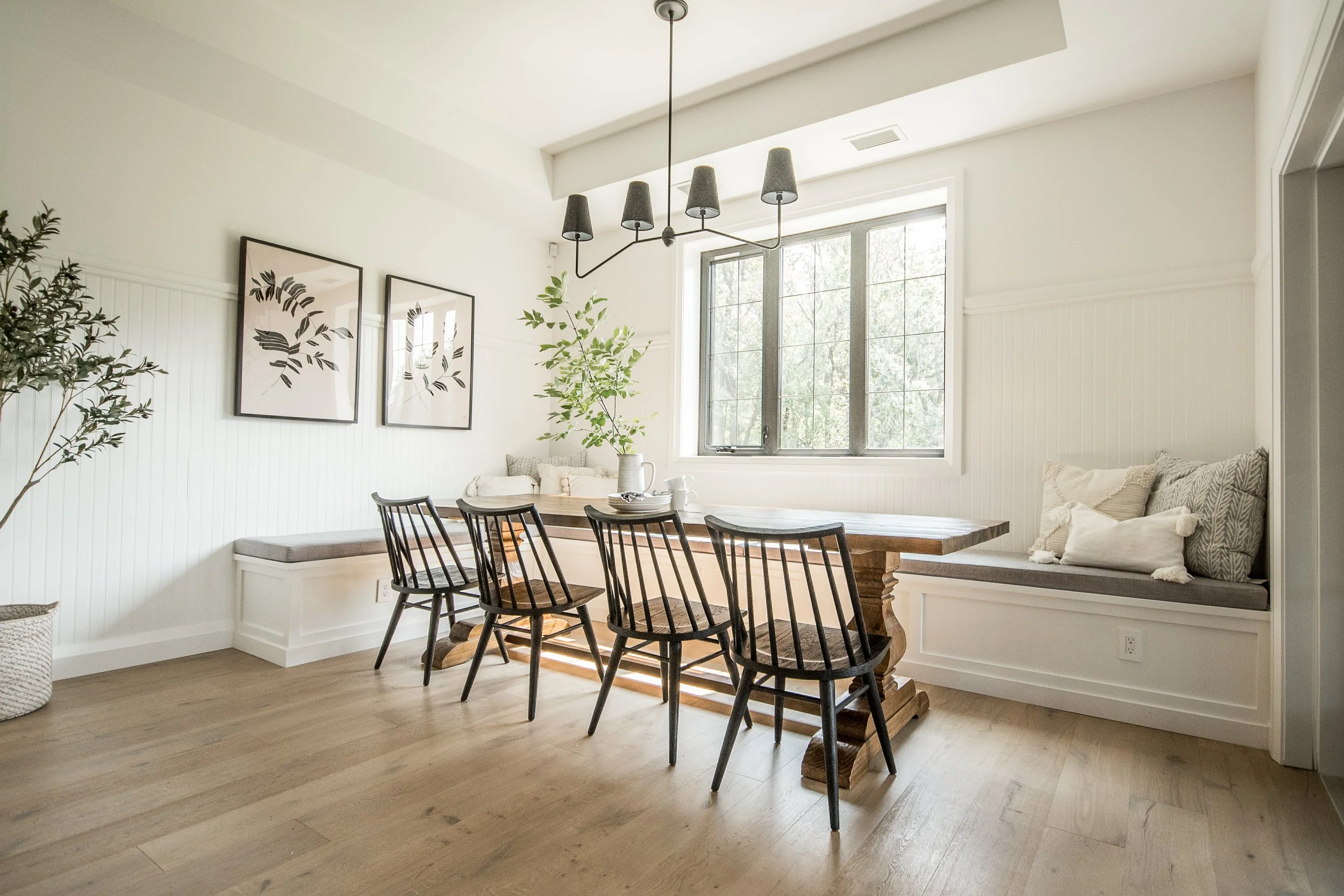 A bright dining area with wood flooring, a wooden table, black chairs, a bench with cushions, wall art, and a large window with trees outside.