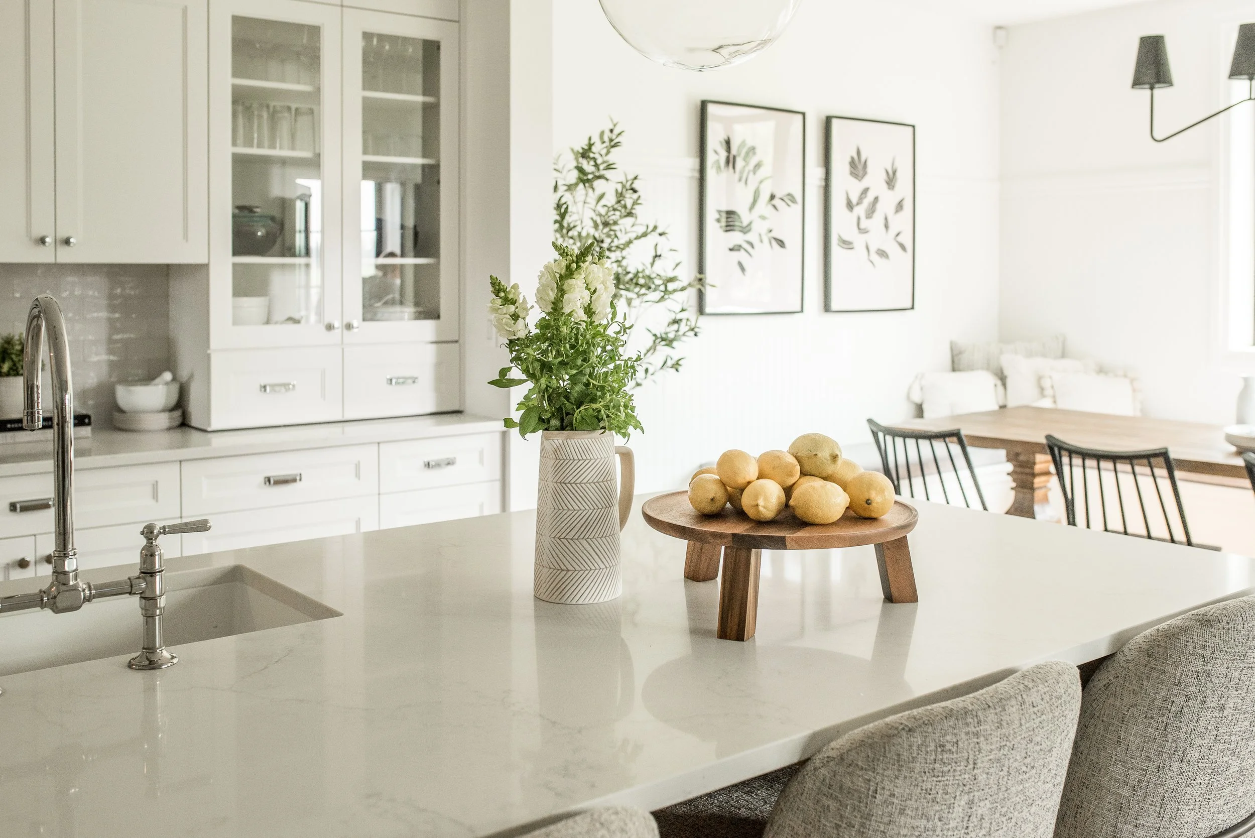 A modern kitchen with white cabinetry, a marble island with a sink, a vase of white flowers, a wooden platform with lemons, black dining chairs, and framed botanical artwork on the wall.