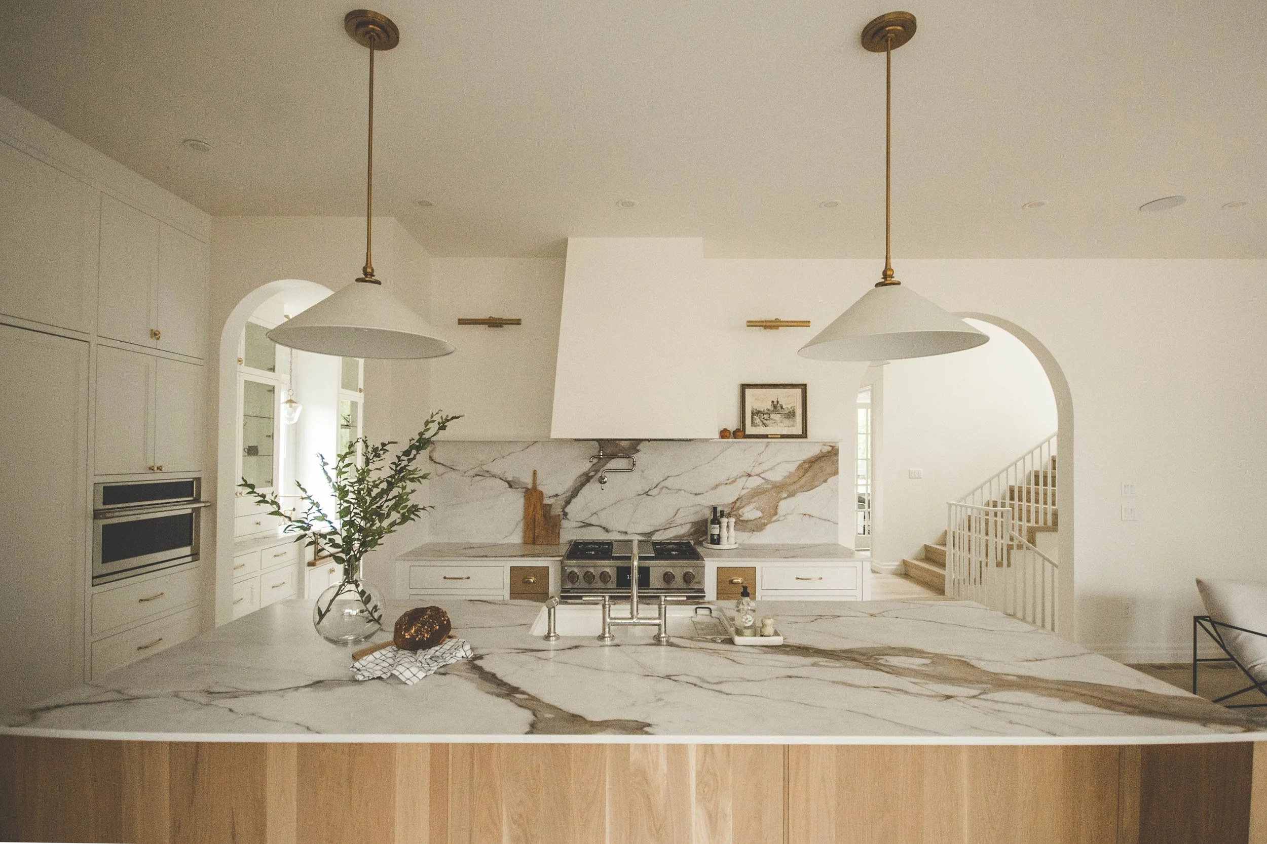 Modern kitchen with marble island and backsplash, white cabinets, and gold accents, illuminated by two pendant lights.