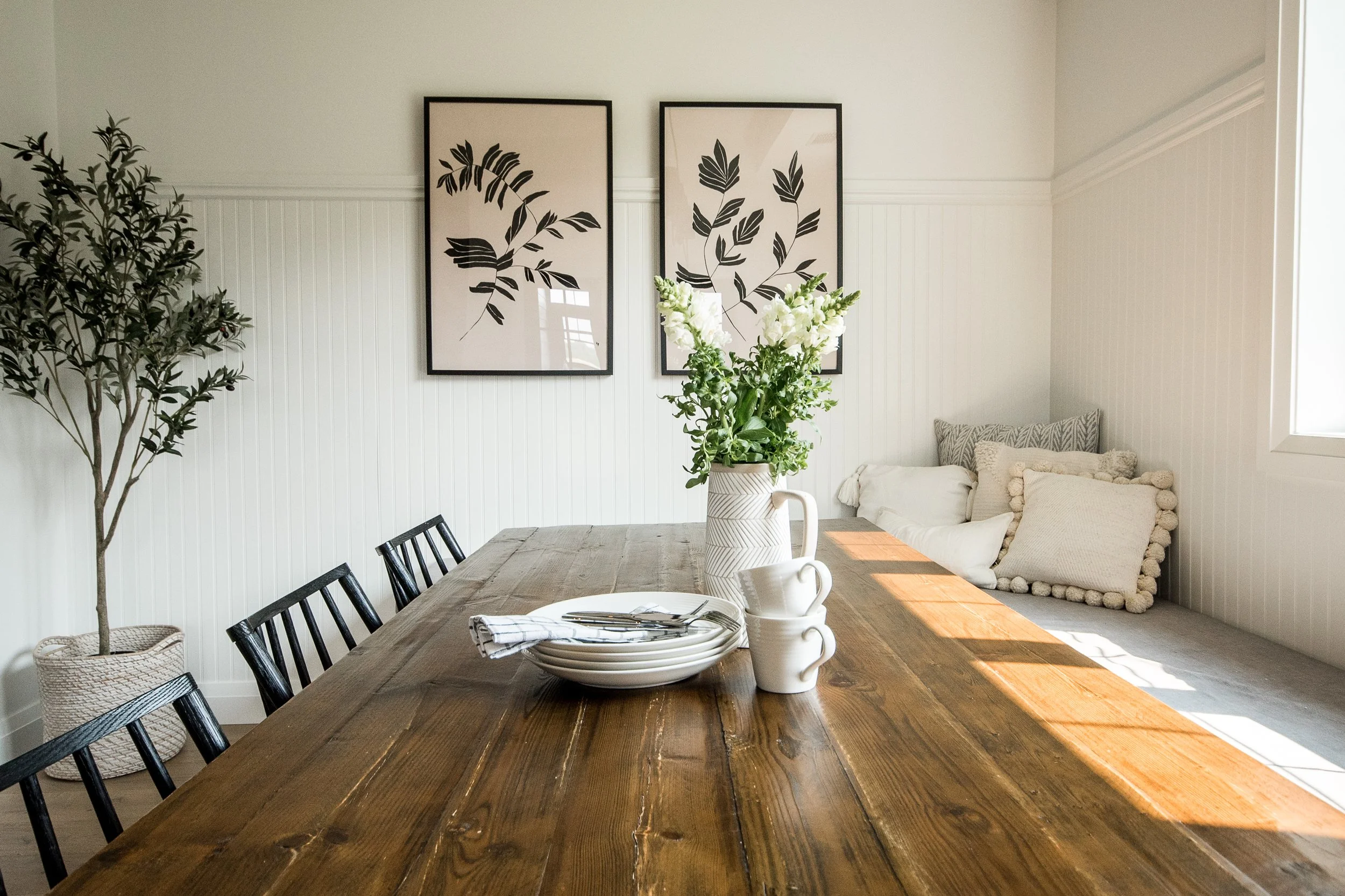 Dining room with a wooden table set with white plates, bowls, and cups, a white pitcher with flowers, black chairs, a potted tree, wall art with black leaf prints, and a window with sunlight.