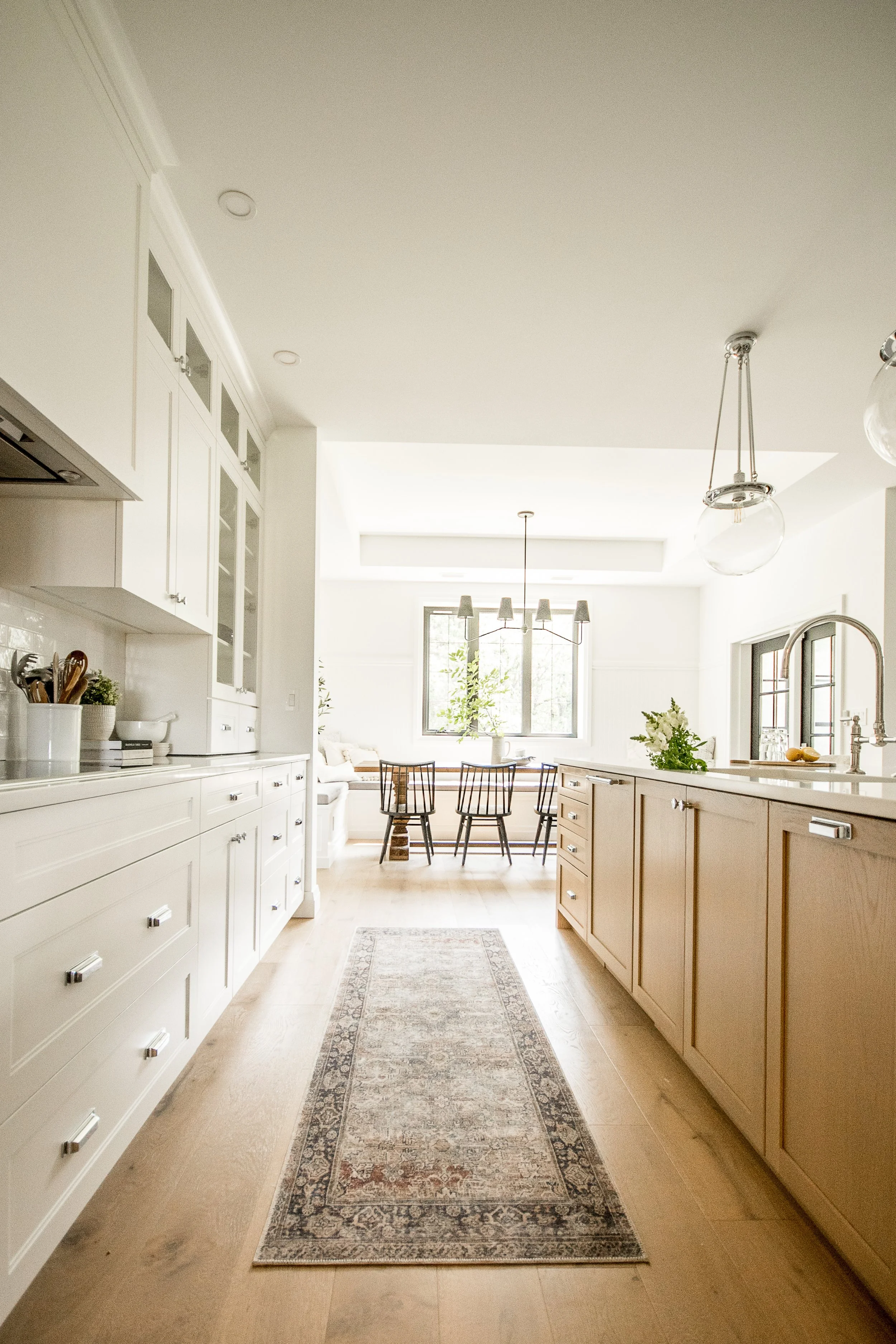 Bright, airy kitchen with white cabinets, a beige island, a small rug on light wood flooring, and a dining area with a table and chairs near large windows.