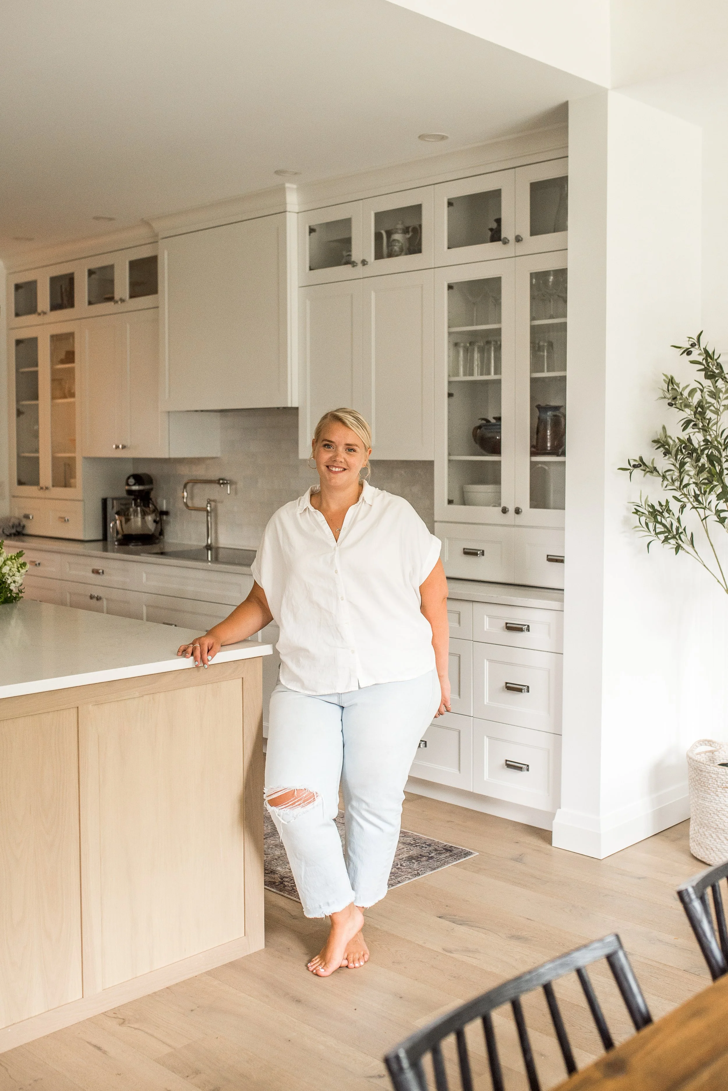 A woman with blonde hair wearing a white shirt and ripped light jeans standing barefooting in a bright kitchen.