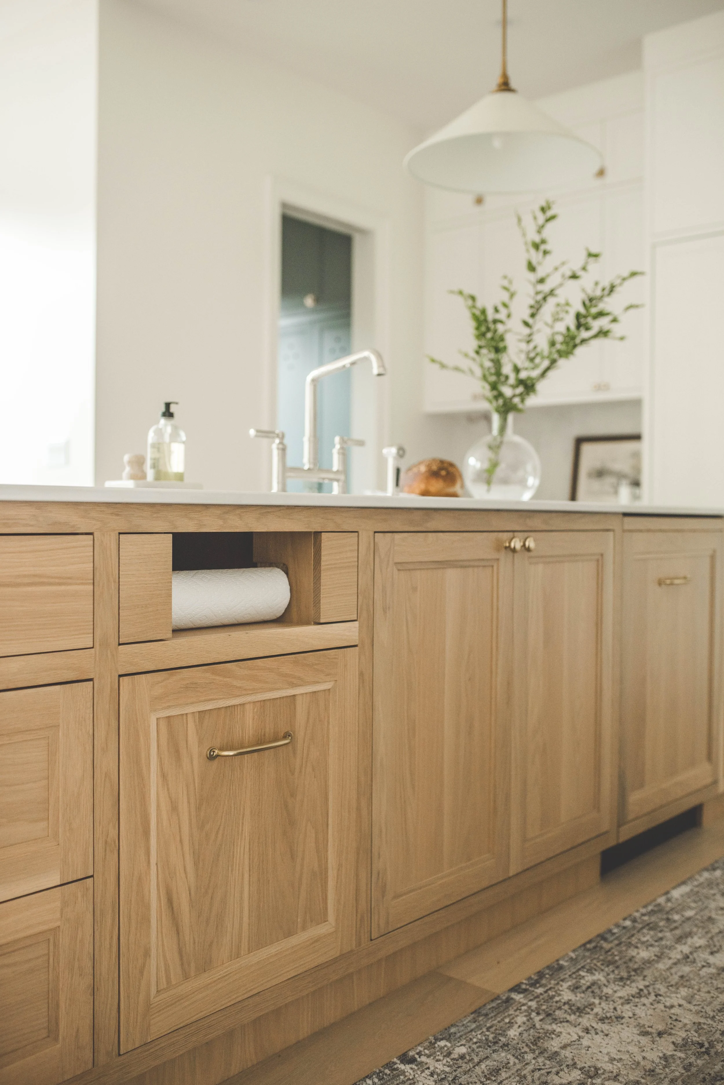 A wooden kitchen countertop with cabinets, a paper towel roll in a niche, a silver faucet, a bottle of hand soap, and a bowl with a loaf of bread. A vase with green foliage is on the countertop, with a framed picture and other decor in the background
