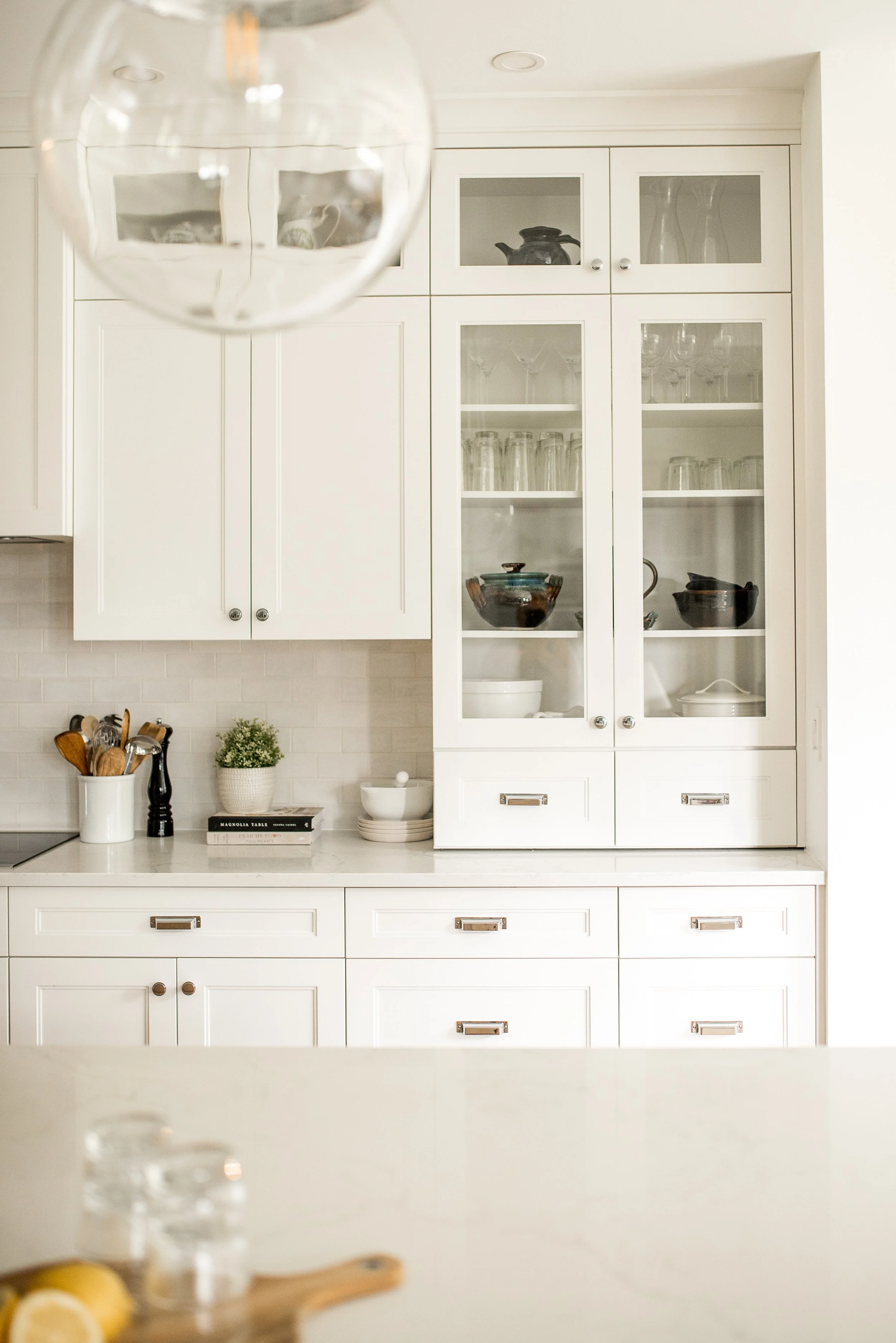 A white kitchen with a glass-front cabinet filled with glassware, bowls, and decorative items, and a countertop with kitchen utensils, a plant, and dishes.