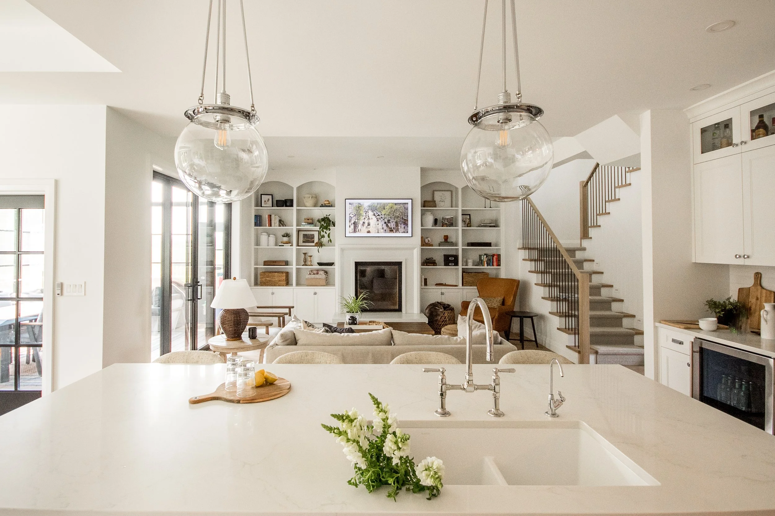 Bright and modern open-concept living room and kitchen with white cabinetry, built-in shelves, a fireplace, and a staircase, decorated with plants and cozy furniture.
