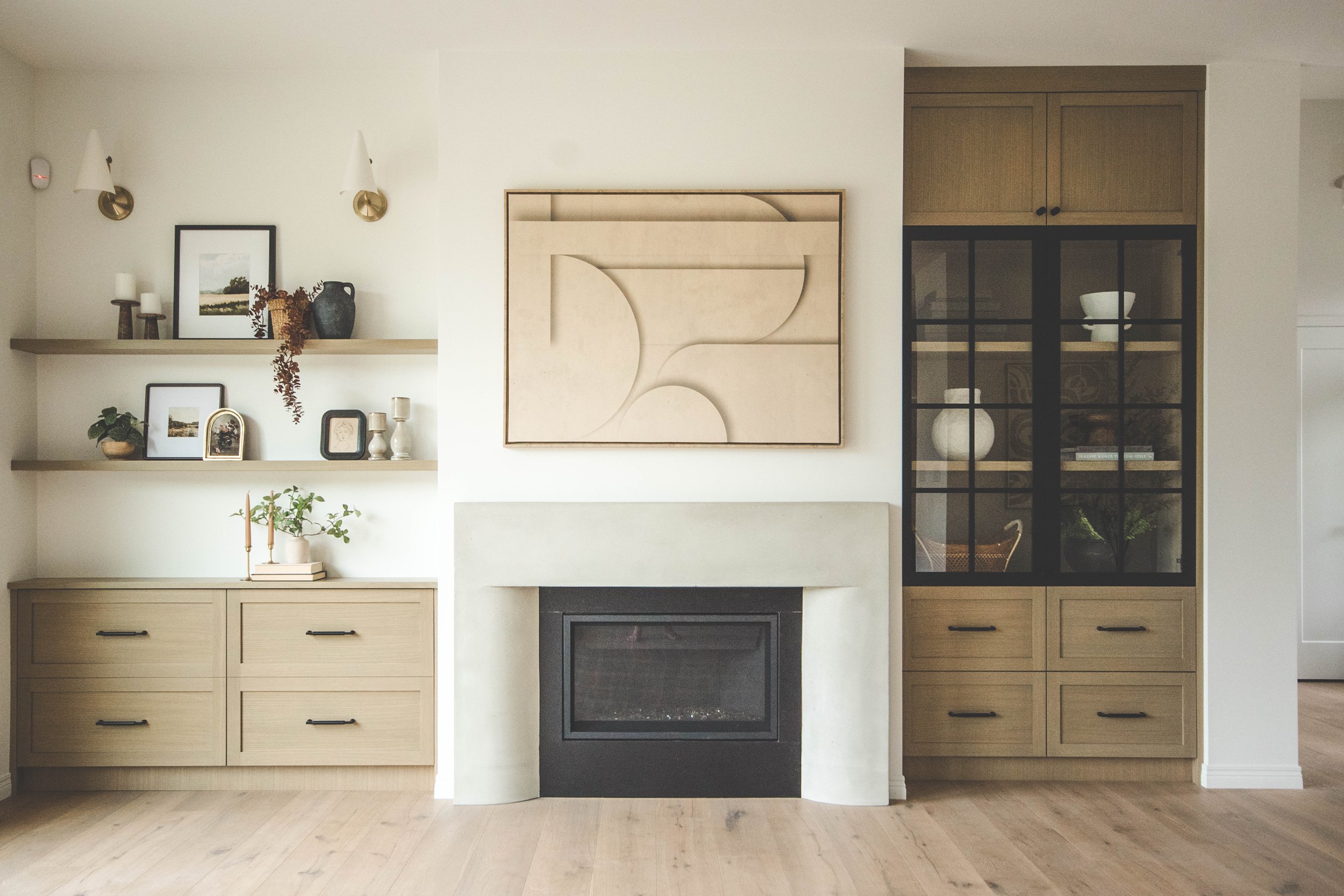 Living room with a fireplace, two wooden cabinets, shelves with decorative items, and a framed abstract art piece above the fireplace.