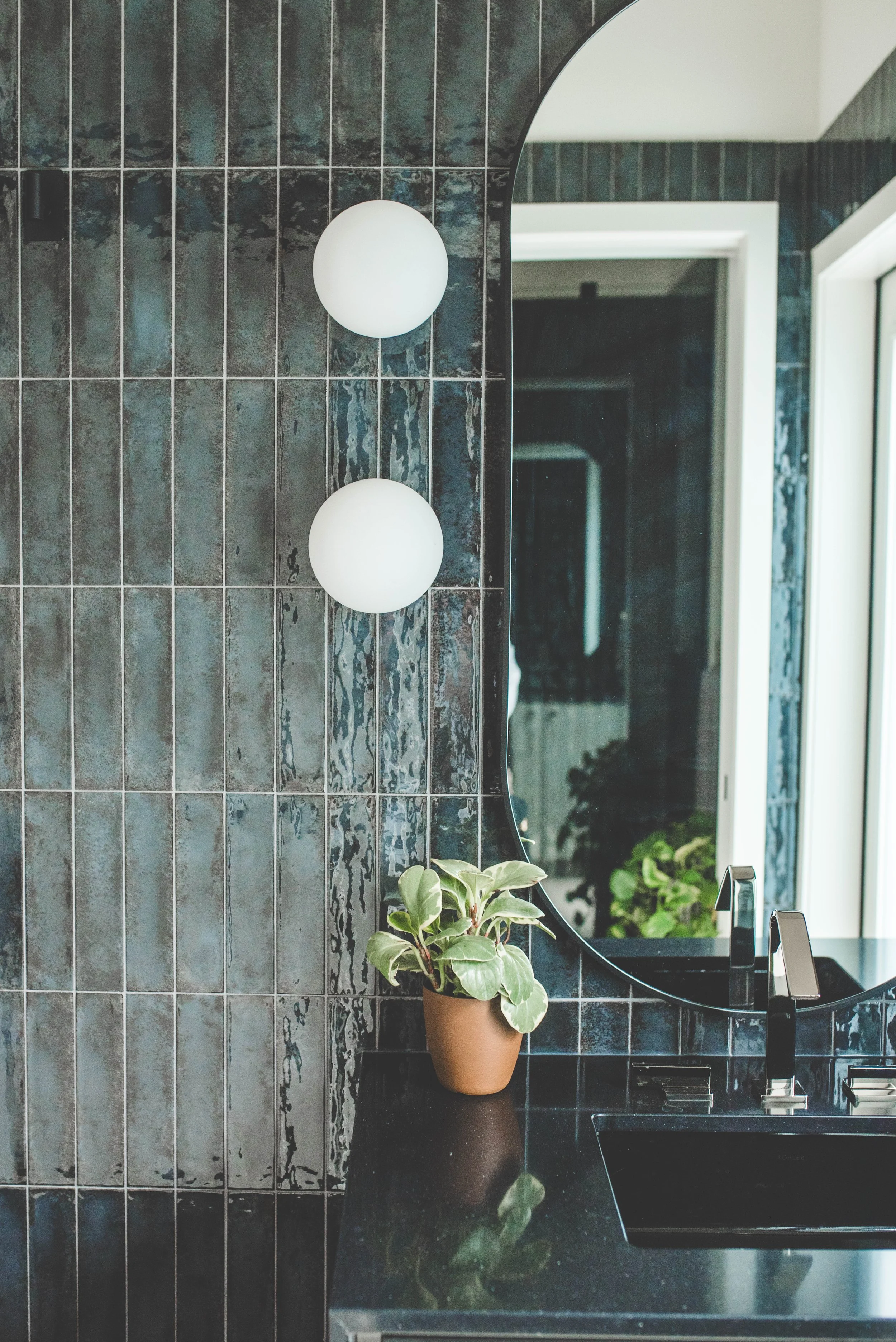 A modern bathroom featuring dark-tiling on the wall, a black countertop with a sink, a potted plant on the counter, and a large mirror with an arched top reflecting part of the room and window.