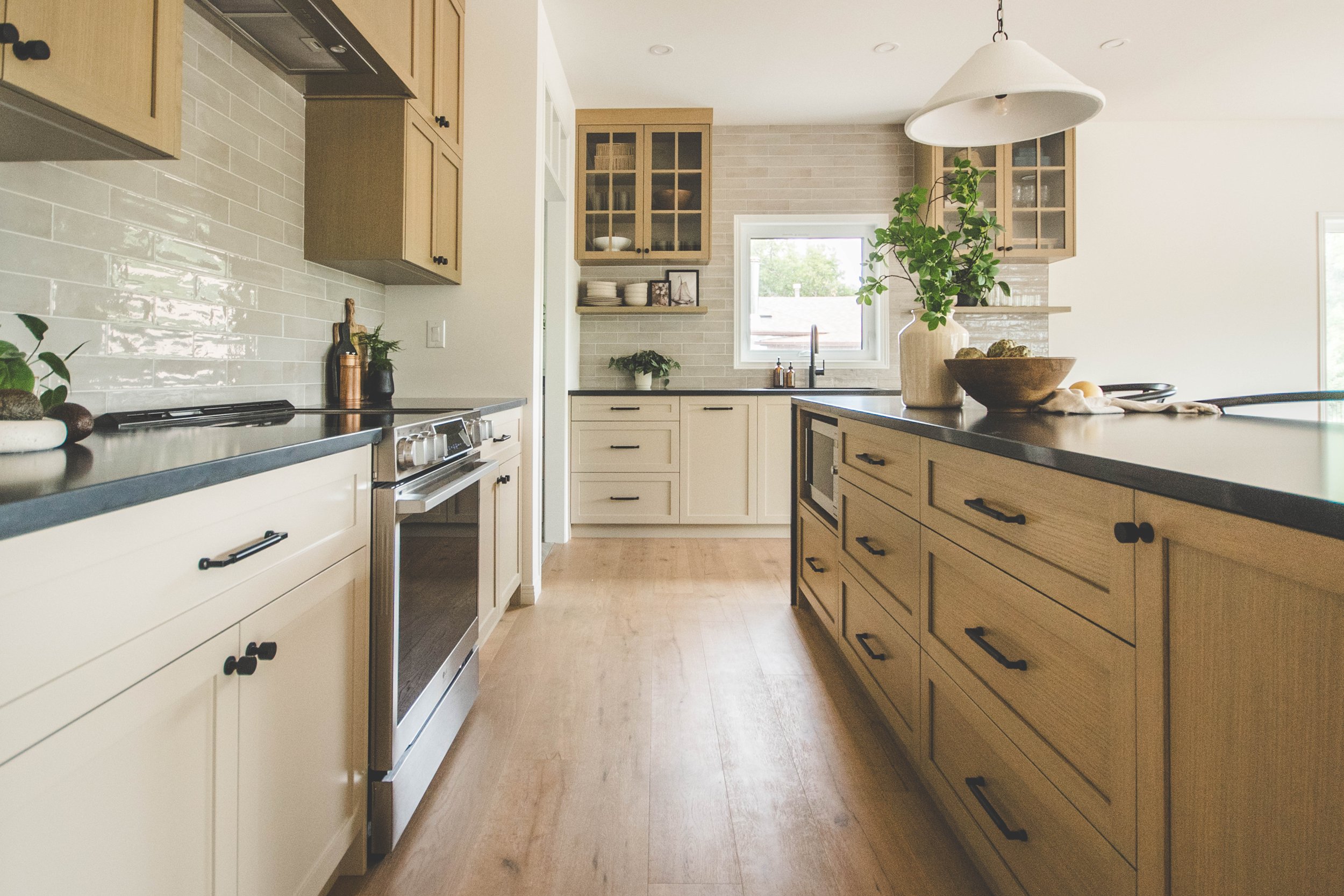 A modern kitchen with light wood cabinets, black countertops, a stainless steel oven, a white subway tile backsplash, and a window above the sink with a green plant on the counter.