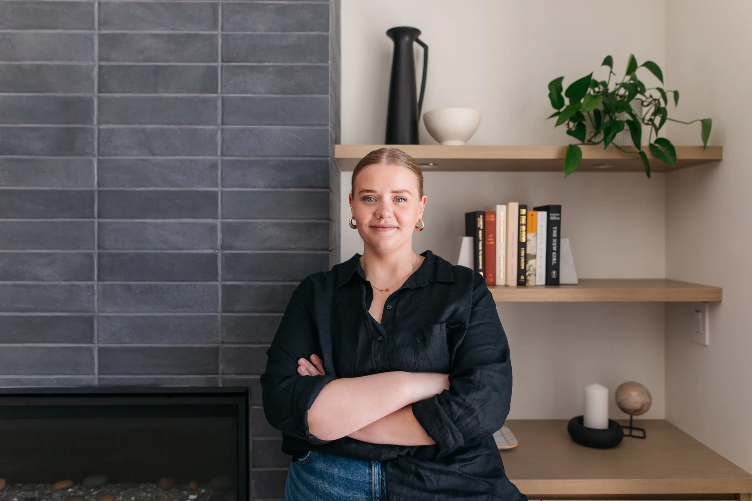 Zoe Henry, lead designer, i wearing a black shirt and gold jewelry, standing with arms crossed in front of a modern living room with a dark grey brick wall, wooden shelves with books, a black vase, a white bowl, a green plant, and decorative objects.