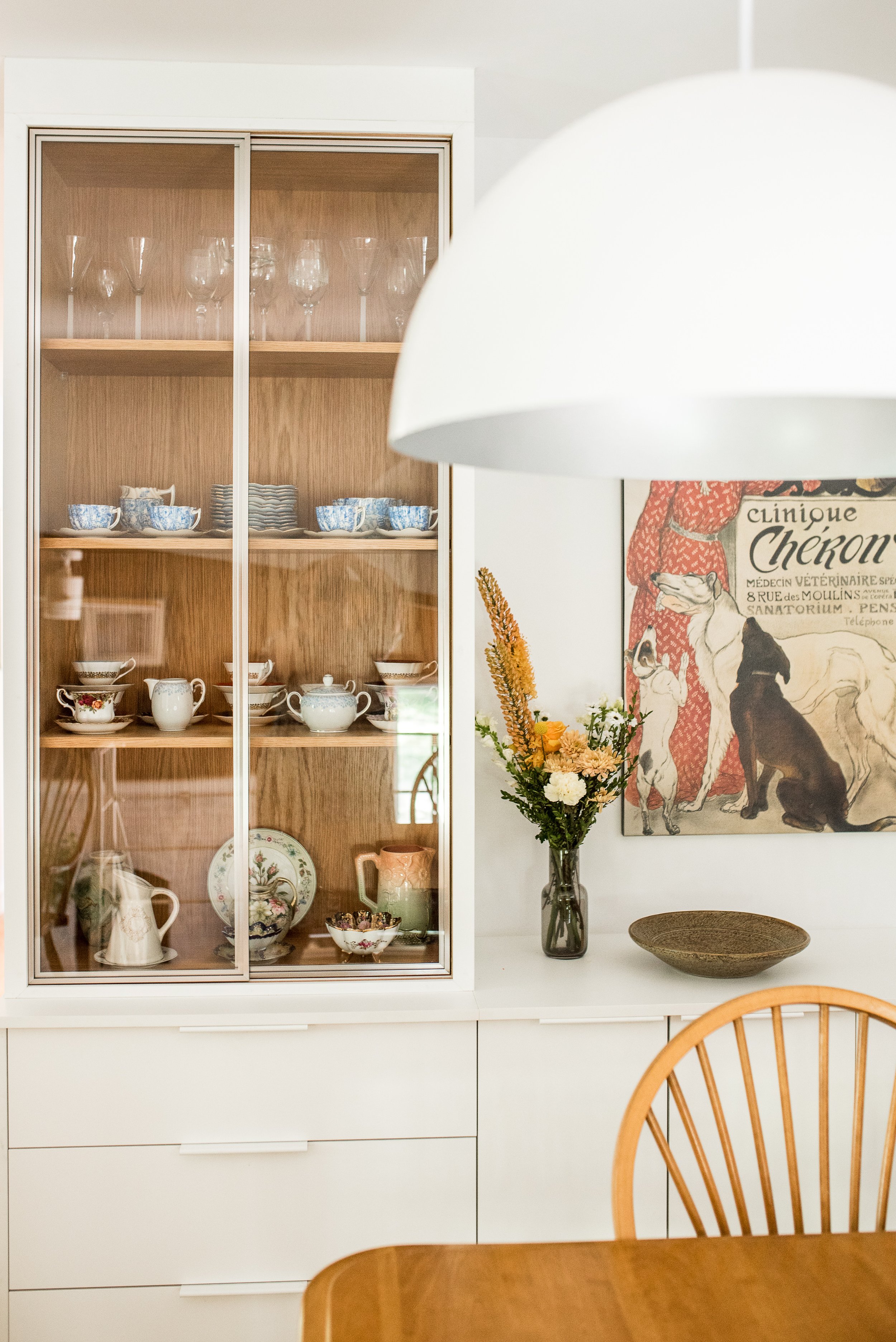 A china cabinet filled with teacups, teapots, and glassware, a vase with flowers, a vintage dog poster, and a wooden chair in a dining area.