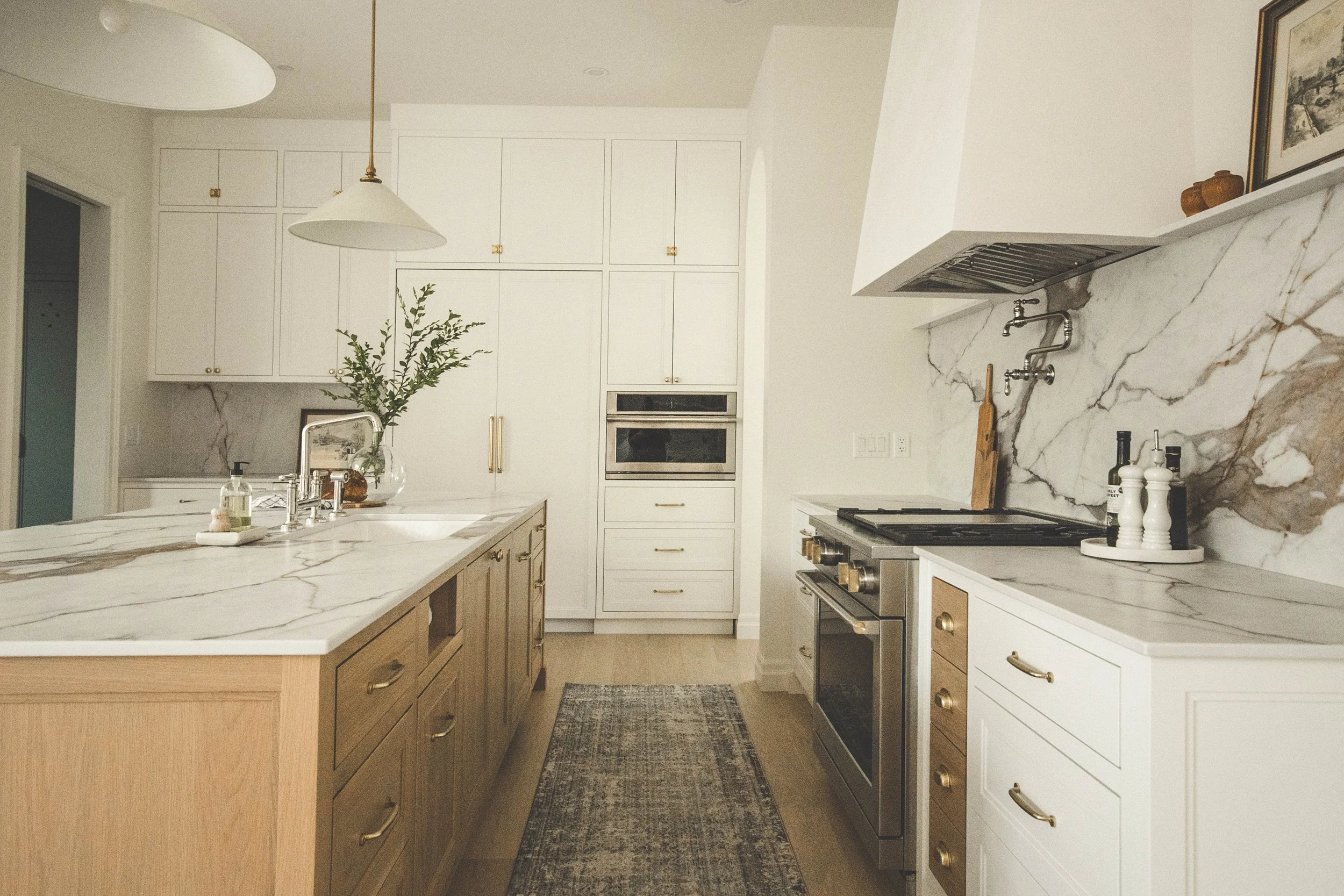 A modern kitchen with white and wood cabinetry, marble countertops and backsplash, a stainless steel stove, and a kitchen island with a large glass vase containing greenery.