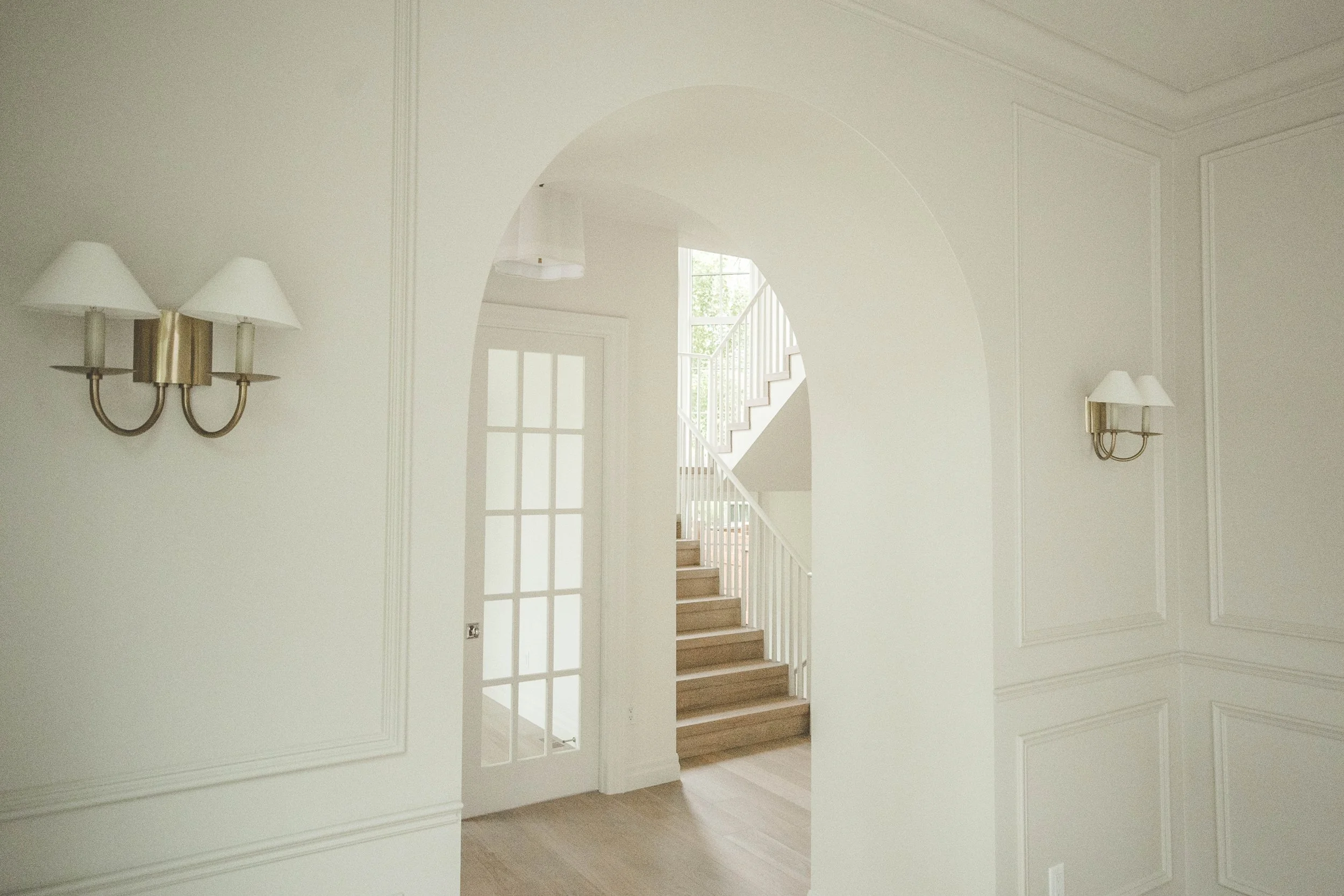 Interior view of a light-colored room with wall-mounted lamps, an arched doorway, and a staircase with white railings and wooden steps leading upward.