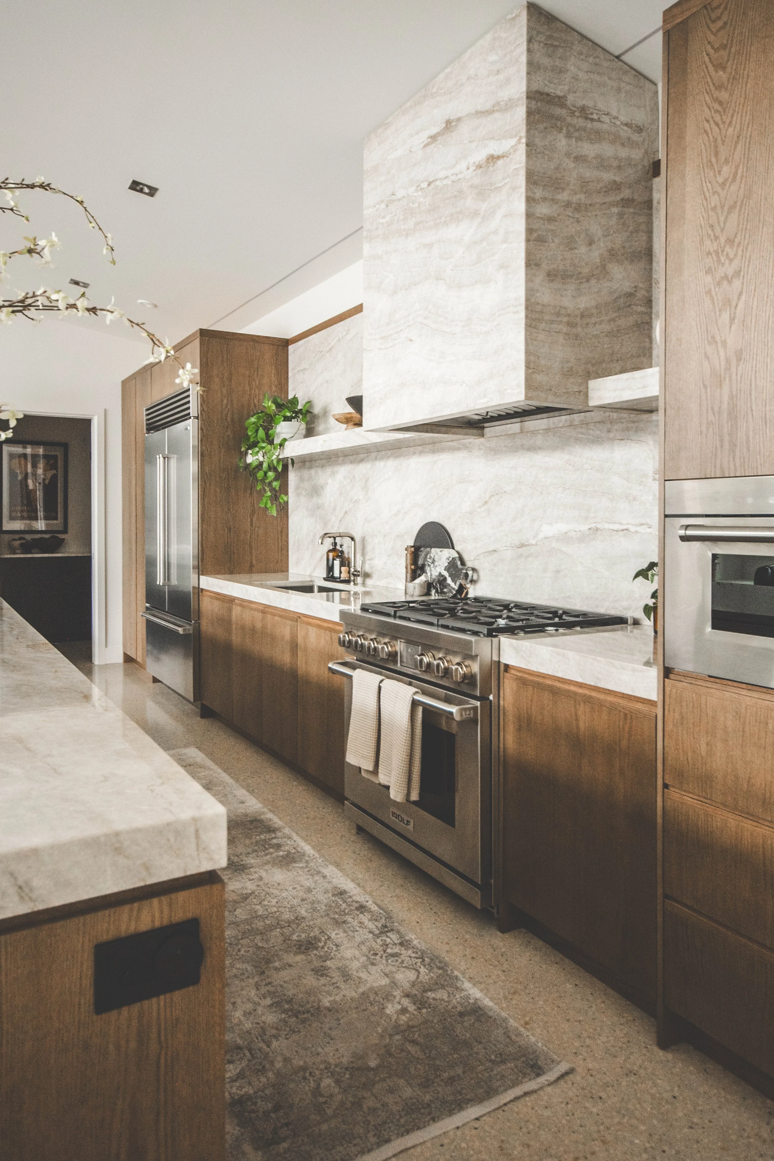 Modern kitchen with wooden cabinets, a stone backsplash, stainless steel appliances including a refrigerator, oven, and microwave, and a countertop with a small plant and kitchen items.