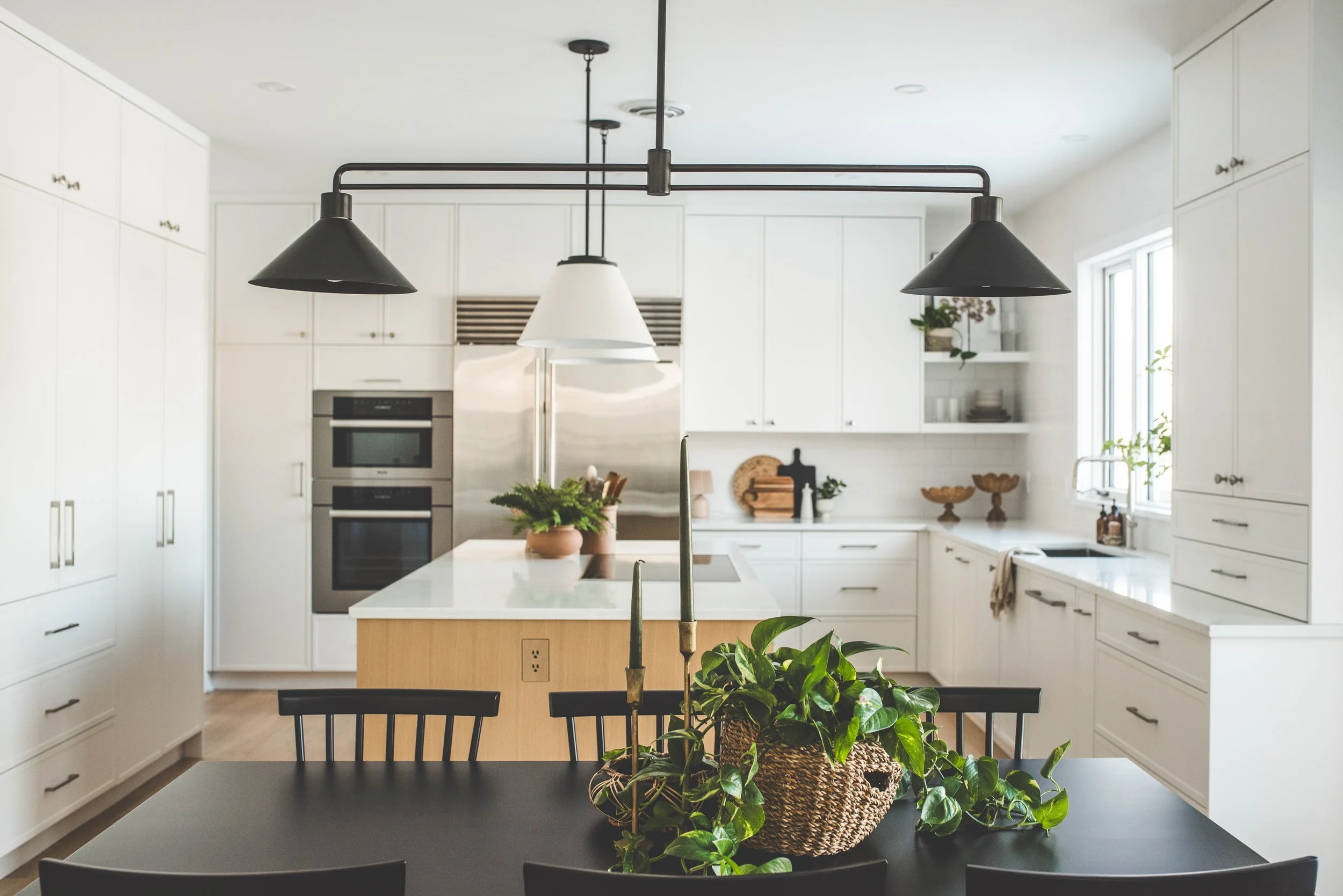 Bright white modern kitchen with black pendant lights, a large island, and a dining table with a plant centerpiece.