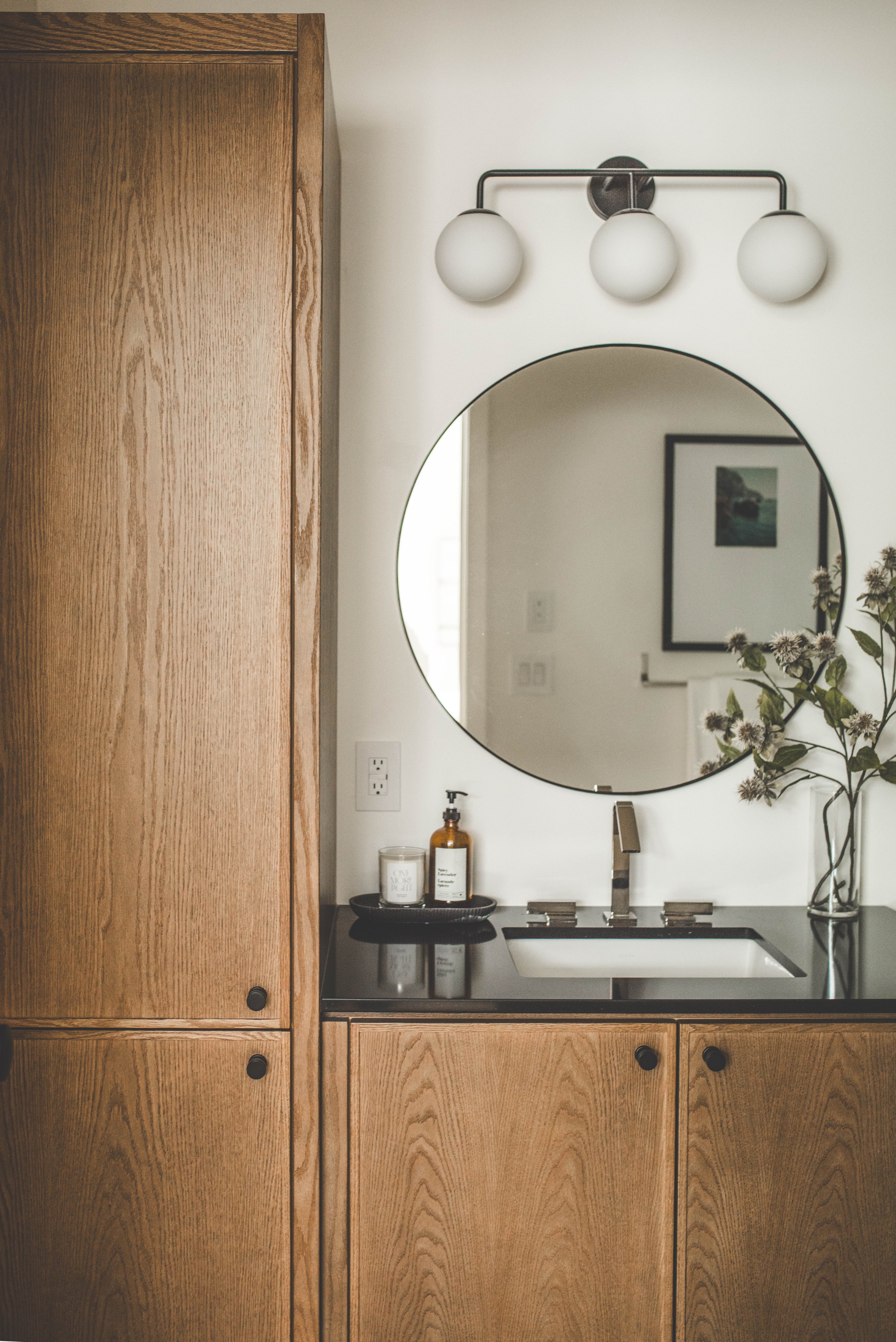 A bathroom vanity with wooden cabinets, a black countertop with a sink, a round mirror, a three-bulb light fixture, a soap dispenser, a candle, and a vase with flowers.