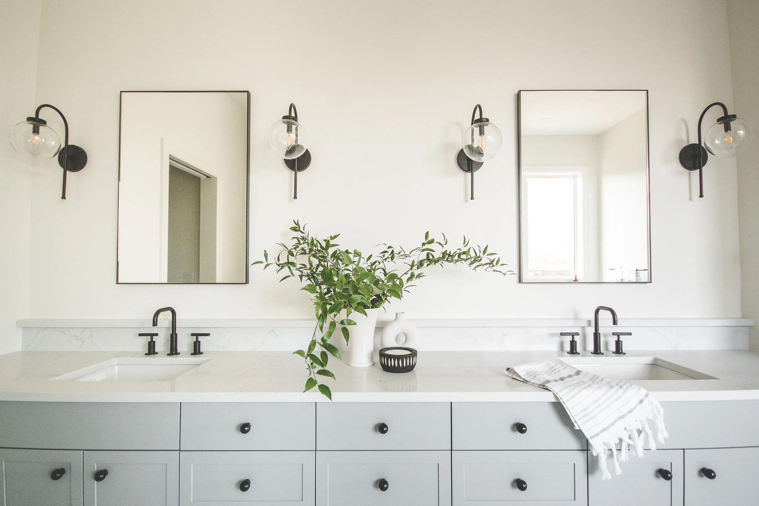 Modern bathroom with dual sinks, black fixtures, two large mirrors, wall-mounted light fixtures, a green plant, vases, and a striped cloth on the countertop.
