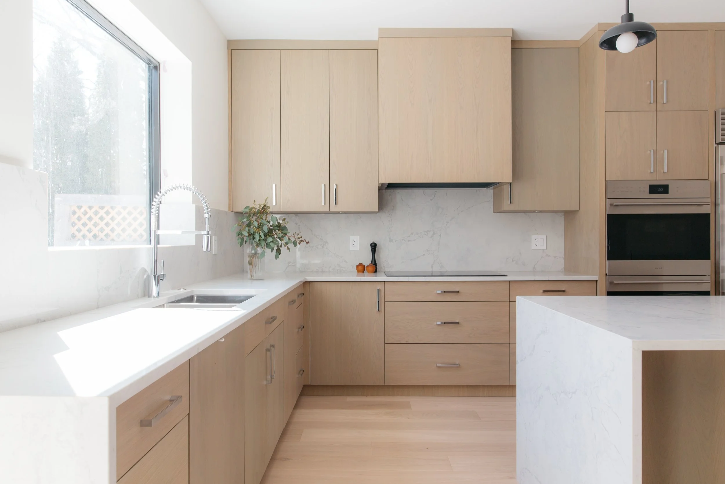 Modern kitchen with light wood cabinets, marble backsplash, white countertops, a stainless steel sink, window, potted plant, and built-in oven.