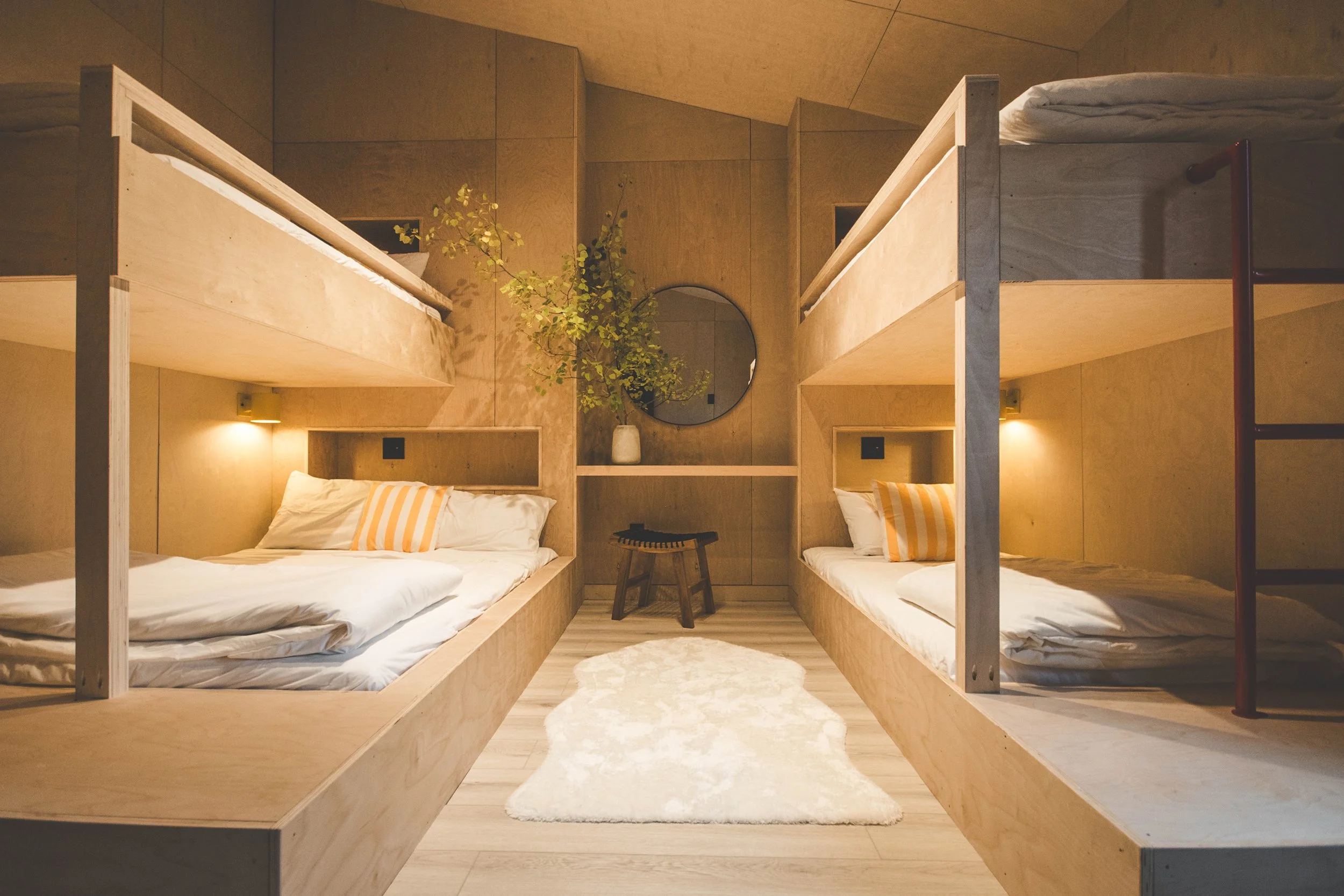 A cozy room with two sets of wooden bunk beds on each side, a round mirror on the back wall, a vase with green foliage, a small plush rug, and a small musical instrument on a stool.