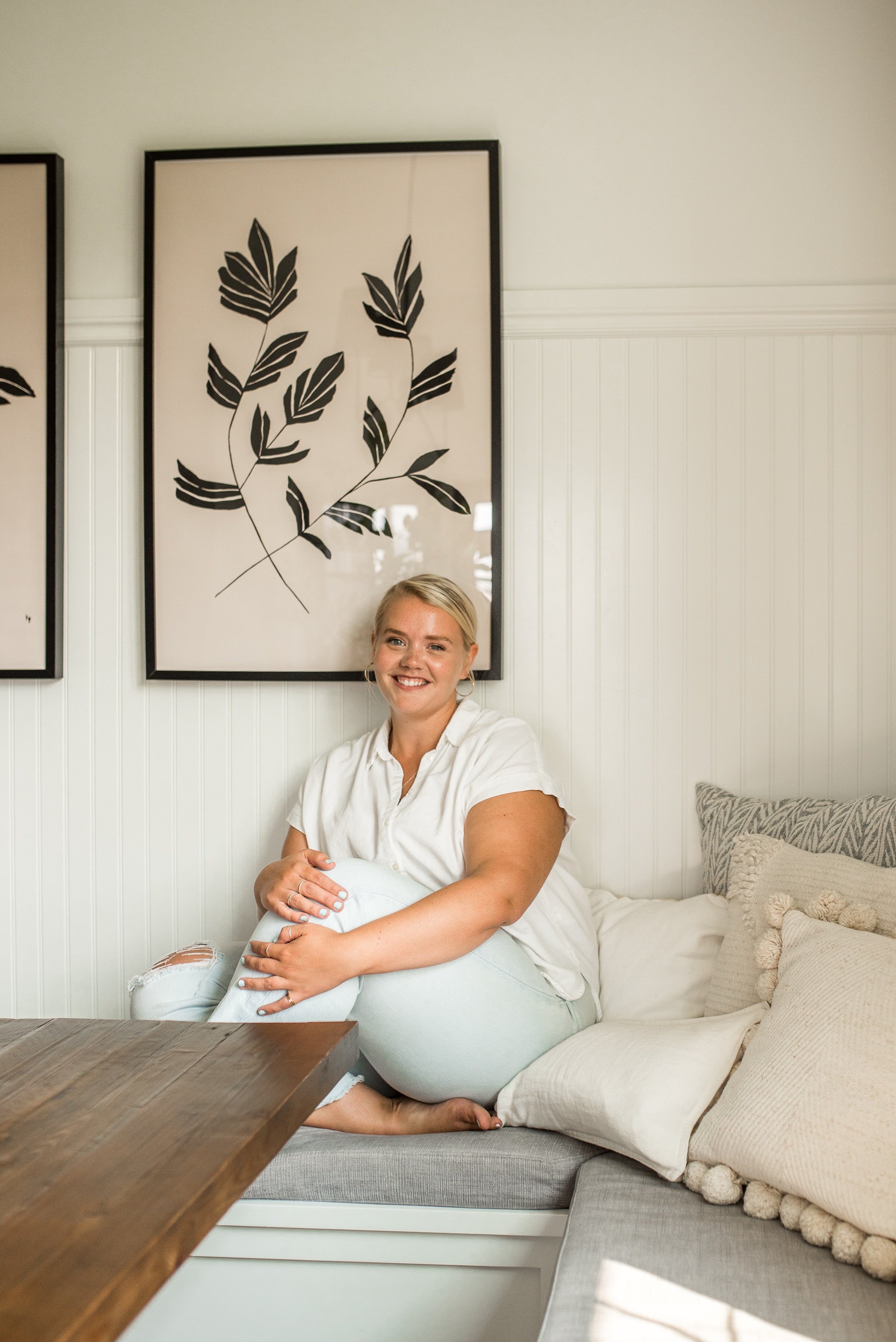 Smiling woman sitting on a cozy built-in window seat, hugging her knee, with framed botanical art on the wall behind her.