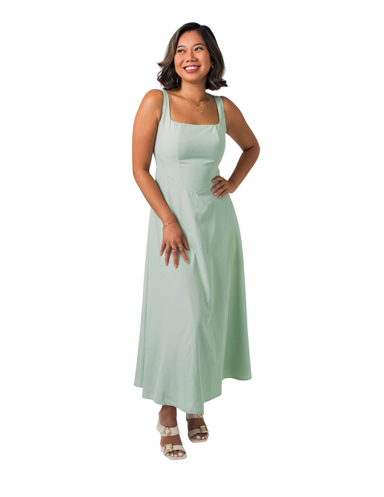 Vanessa Luv - Vancouver BC - Divera Leadership A woman with shoulder-length dark hair, smiling, wearing a sleeveless light green maxi dress, gold jewelry, and white sandals, standing with one hand on her hip.
