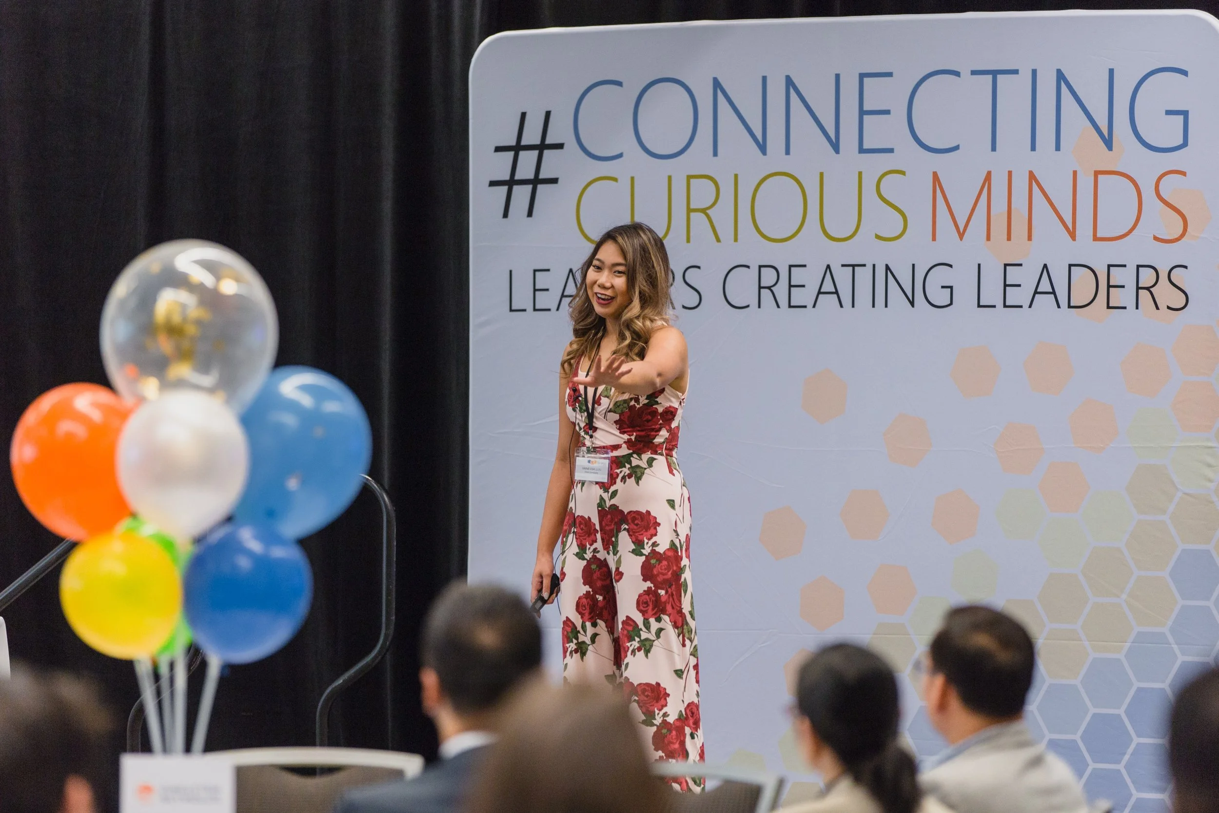 A woman speaking on stage at a conference. She is wearing a floral jumpsuit and standing in front of a backdrop with hashtags and text about connecting, curious minds, and creating leaders. There are balloons in the foreground and some seated attendees visible.