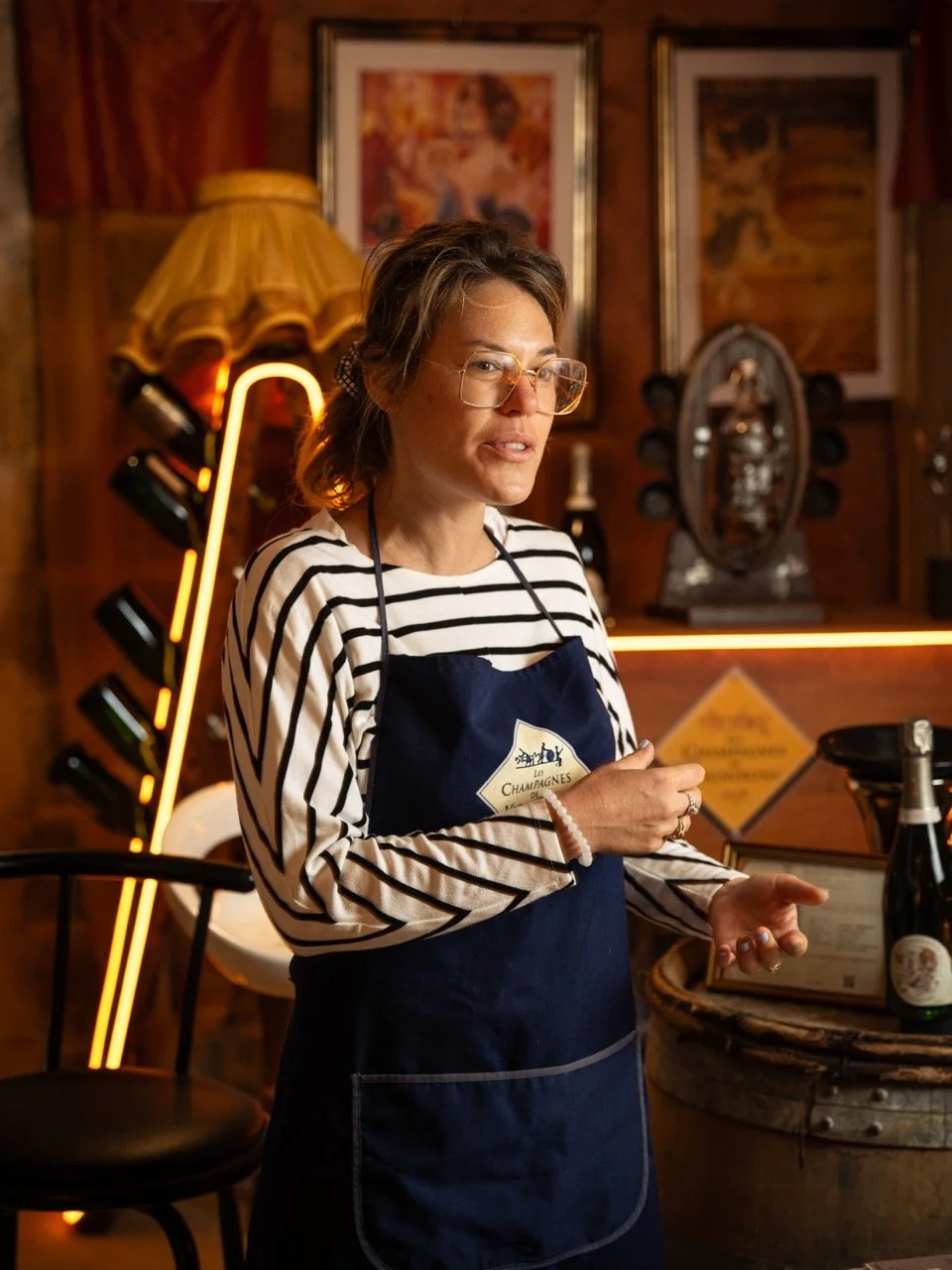 Une vigneronne dans sa cave de dégustation portant un tablier marine et un pull à rayures noires et blanches, avec des lunettes, dans un cadre chaleureux avec des œuvres d'art et une lampe vintage en arrière-plan.