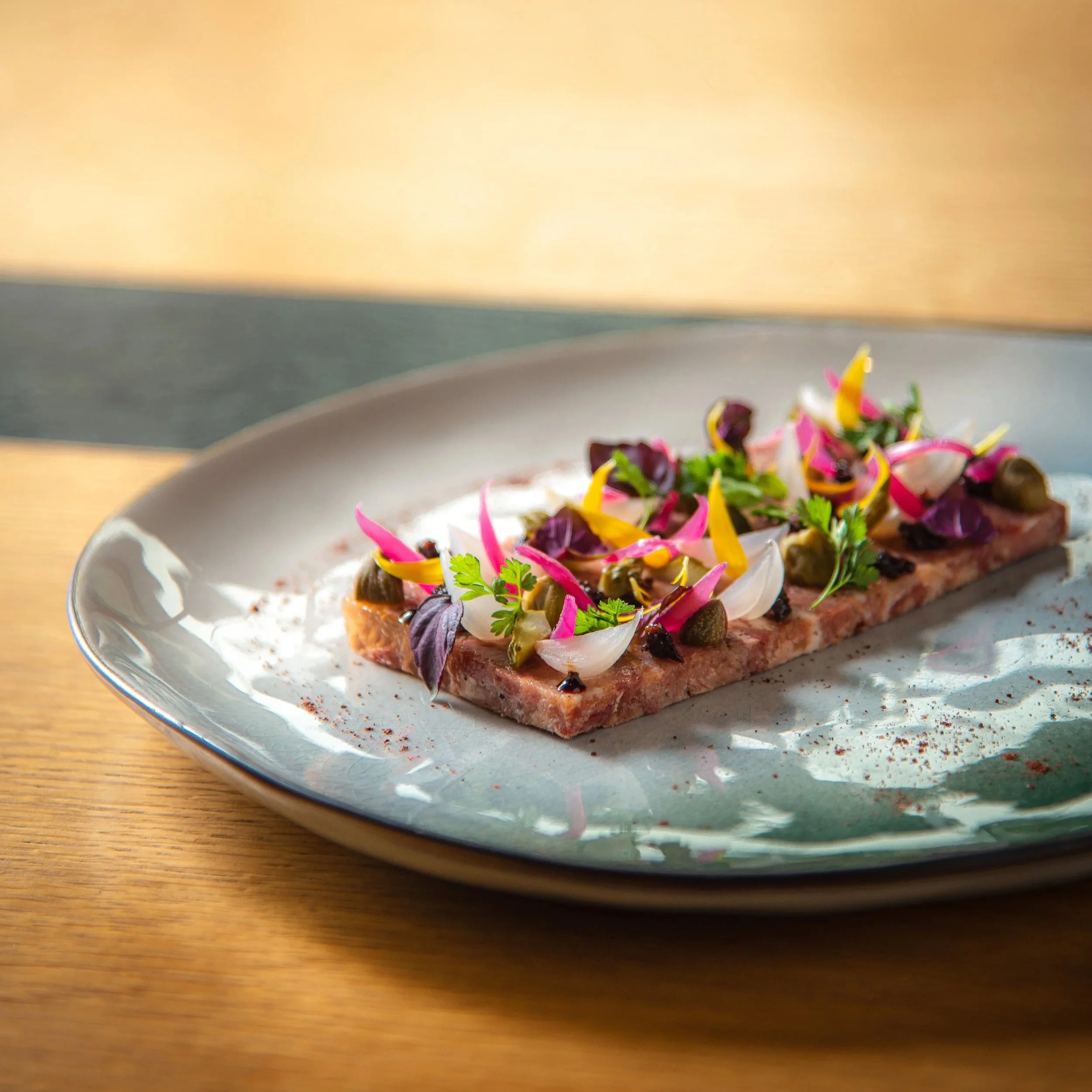 Plat réalisé par le restaurant jardin des crayeres. Une tranche de viande crue garnie de différentes herbes, légumes et condiments colorés, servie sur une assiette. Photo prise par Théo WALLYN, TheWallPhoto.