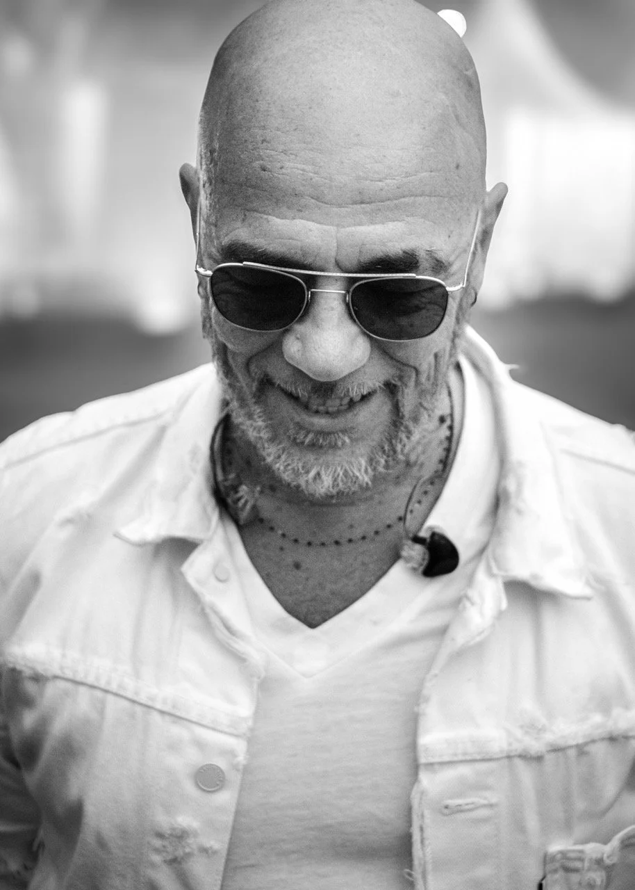 Homme chauve portant des lunettes de soleil, souriant, en chemise blanche avec un micro attaché à son col.