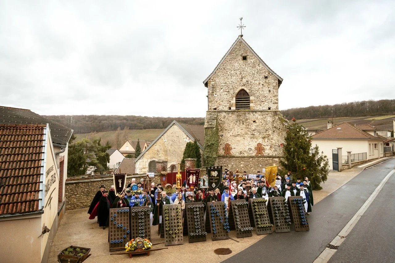 Groupe de personnes en costumes traditionnels lors de la saint Vincent dans une rue en champagne, avec une vieille église en pierre et une colline en arrière-plan.