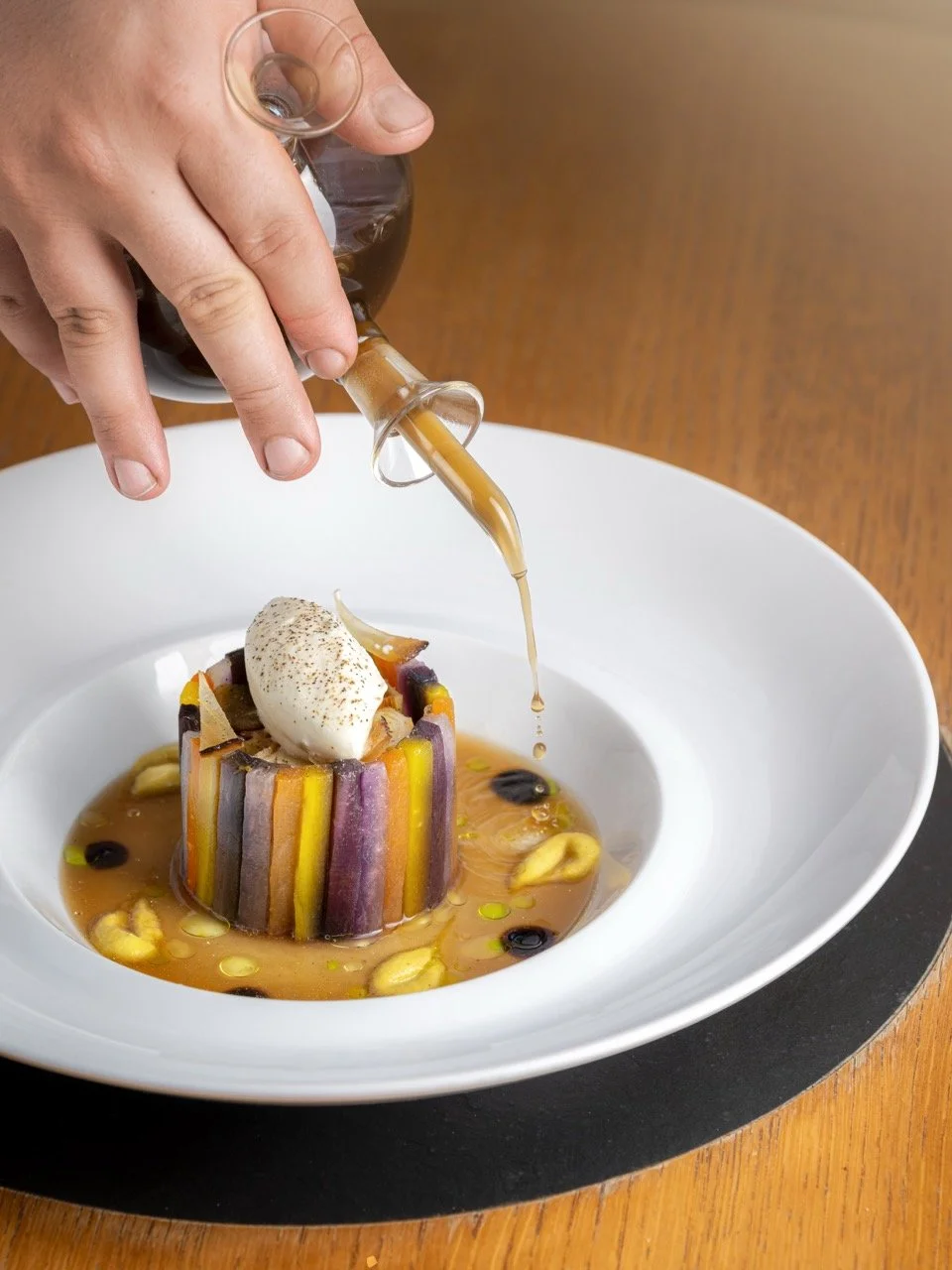 Une personne verse une sauce sur un plat de légumes colorés et une boule de glace dans un bol blanc.Au jardin des crayeres.  Le plat est présenté dans une assiette blanche et repose sur un plateau noir. Photo prise par Théo WALLYN, TheWallPhoto.