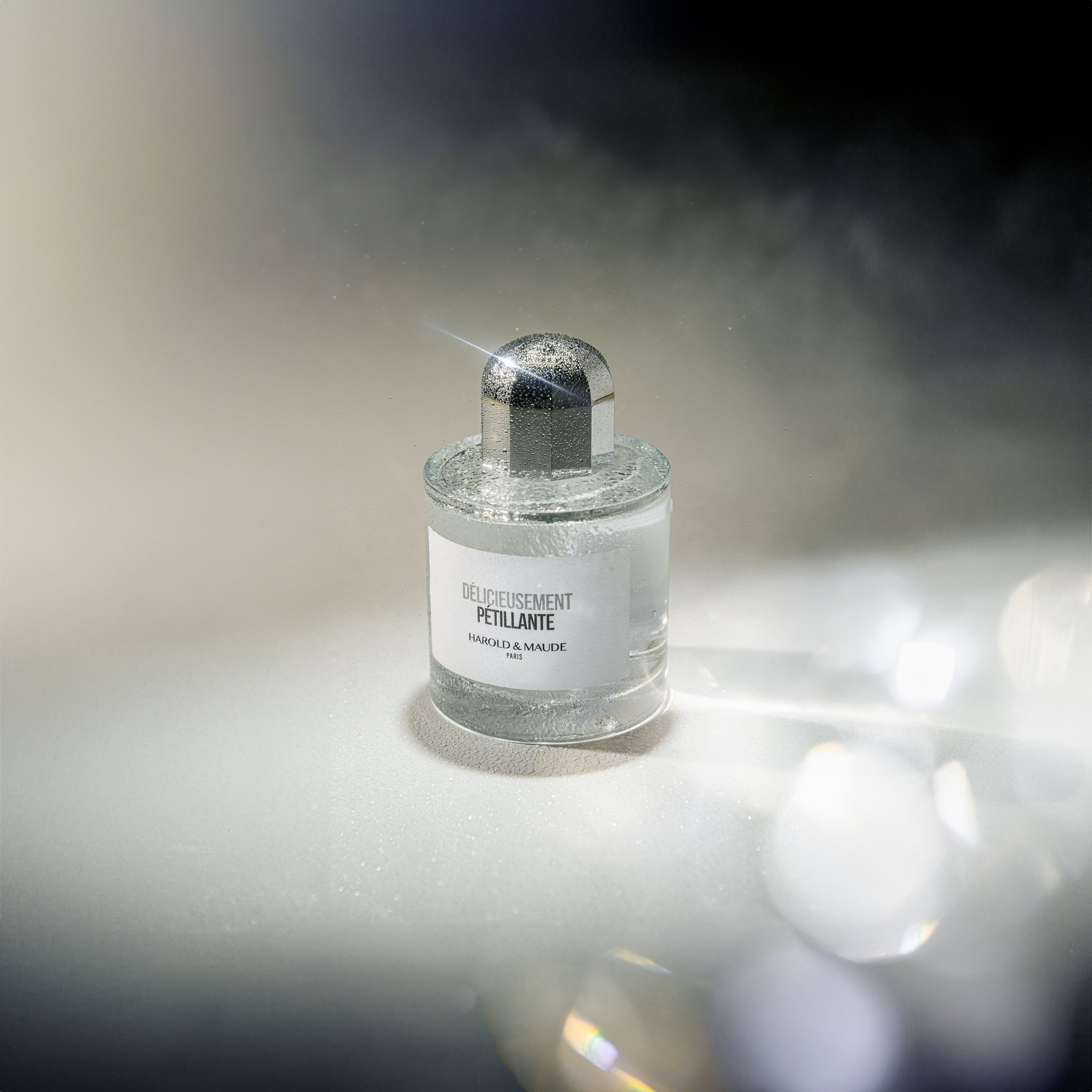 Un flacon de parfum transparent avec une étiquette blanche portant la mention « Délicieusement Pétillante » ainsi que le nom de la marque Harold & Maude, posé sur une surface claire avec des reflets de lumière. Photo prise par Théo WALLYN, TheWallPho