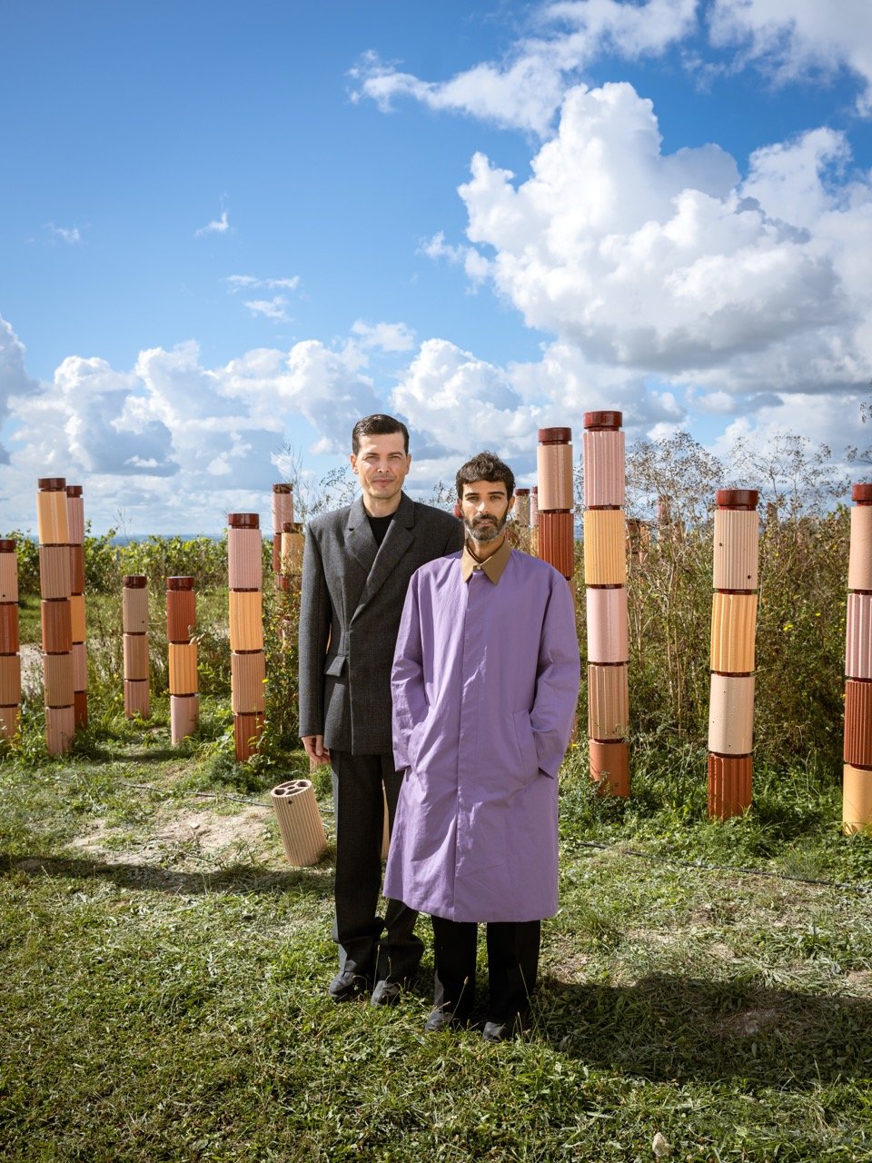 Deux designer debout dans une vigne du champagne Perrier Jouet avec des colonnes en terre cuite décoratives, un ciel bleu avec des nuages blancs, et des buissons derrière eux.