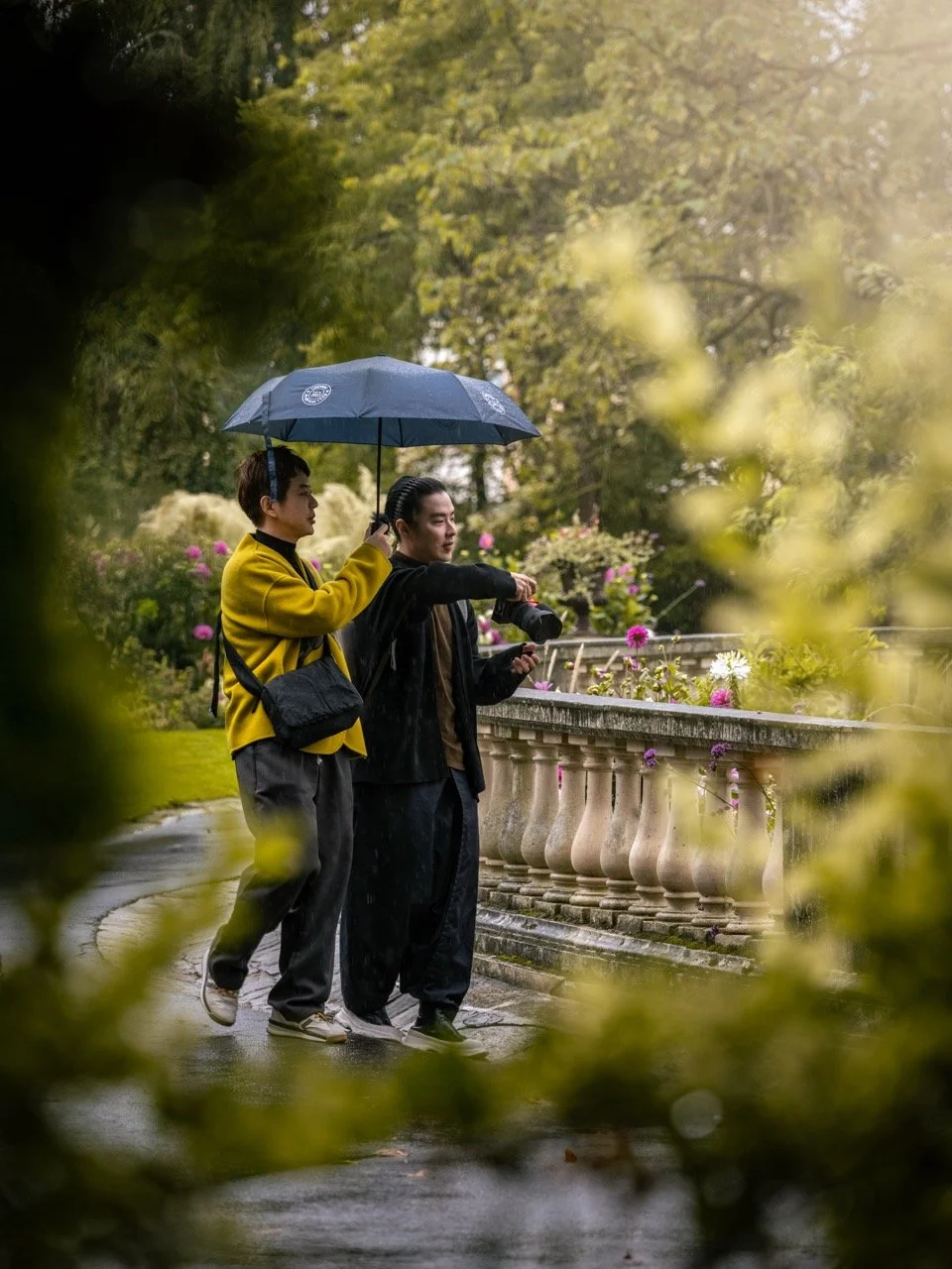 Deux jeunes hommes regardant et prenant des photos dans un jardin sous la pluie, avec un parapluie, entourés de fleurs et d'arbres en arrière-plan.