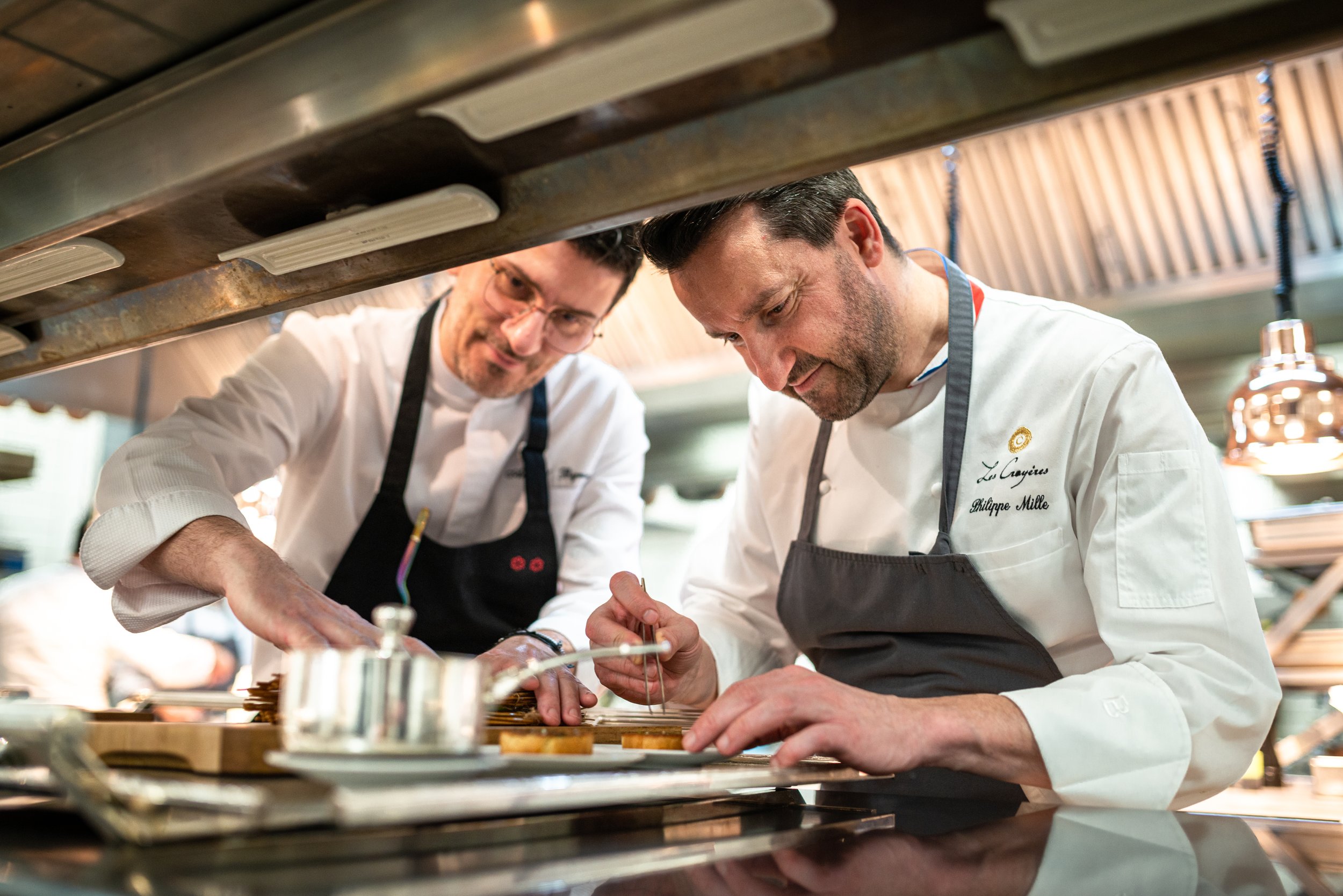Deux chefs en cuisine, dont Phillipe Mille, concentrés sur la préparation de plats, vus de dessous d'une hotte de ventilation. Photo prise par Théo WALLYN, TheWallPhoto.