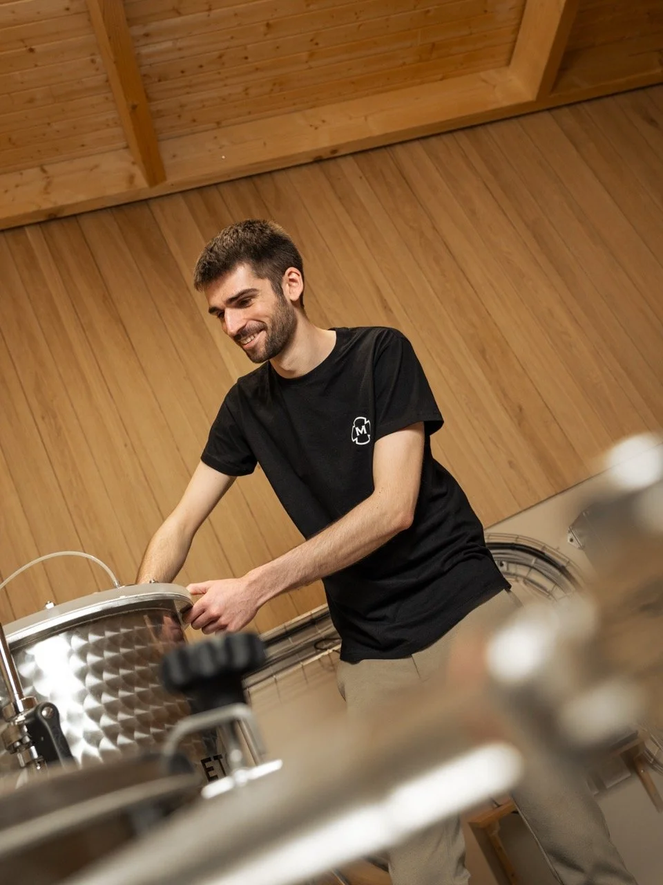 Un homme souriant en t-shirt noir, travaillant dans un environnement en bois, manipulant un équipement métallique, vinaigrerie "les enfants de Bachus". Photo prise par Théo WALLYN, TheWallPhoto.