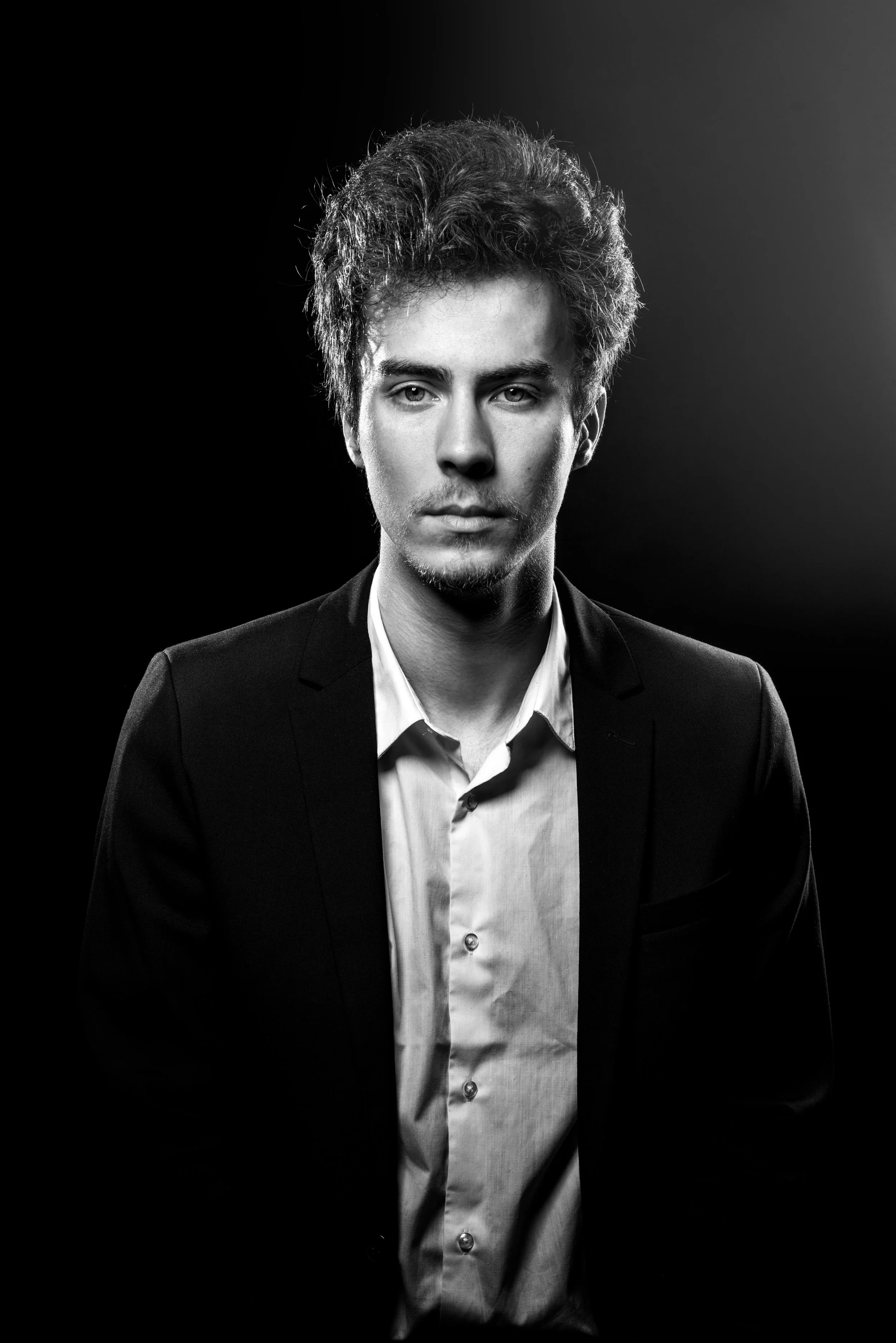 Portrait en noir et blanc d'un jeune homme avec des cheveux bouclés, portant une chemise blanche et une veste noire, posé contre un fond sombre en studio. C'est le portrait d'un acteur. Photo prise par Théo WALLYN, TheWallPhoto.
