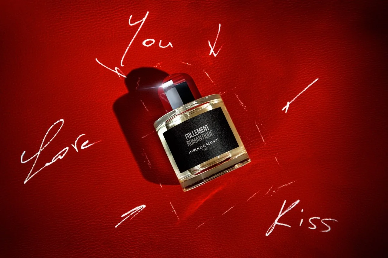 Un flacon de parfum nommé 'FOLLEMENT ROMANTIQUE' de chez Harold et Maude posé sur une surface rouge, entouré de mots manuscrits en blanc comme 'You', 'Love', 'Kiss', avec un jeu de lumière et d'ombres. Photo prise par Théo WALLYN, TheWallPhoto.